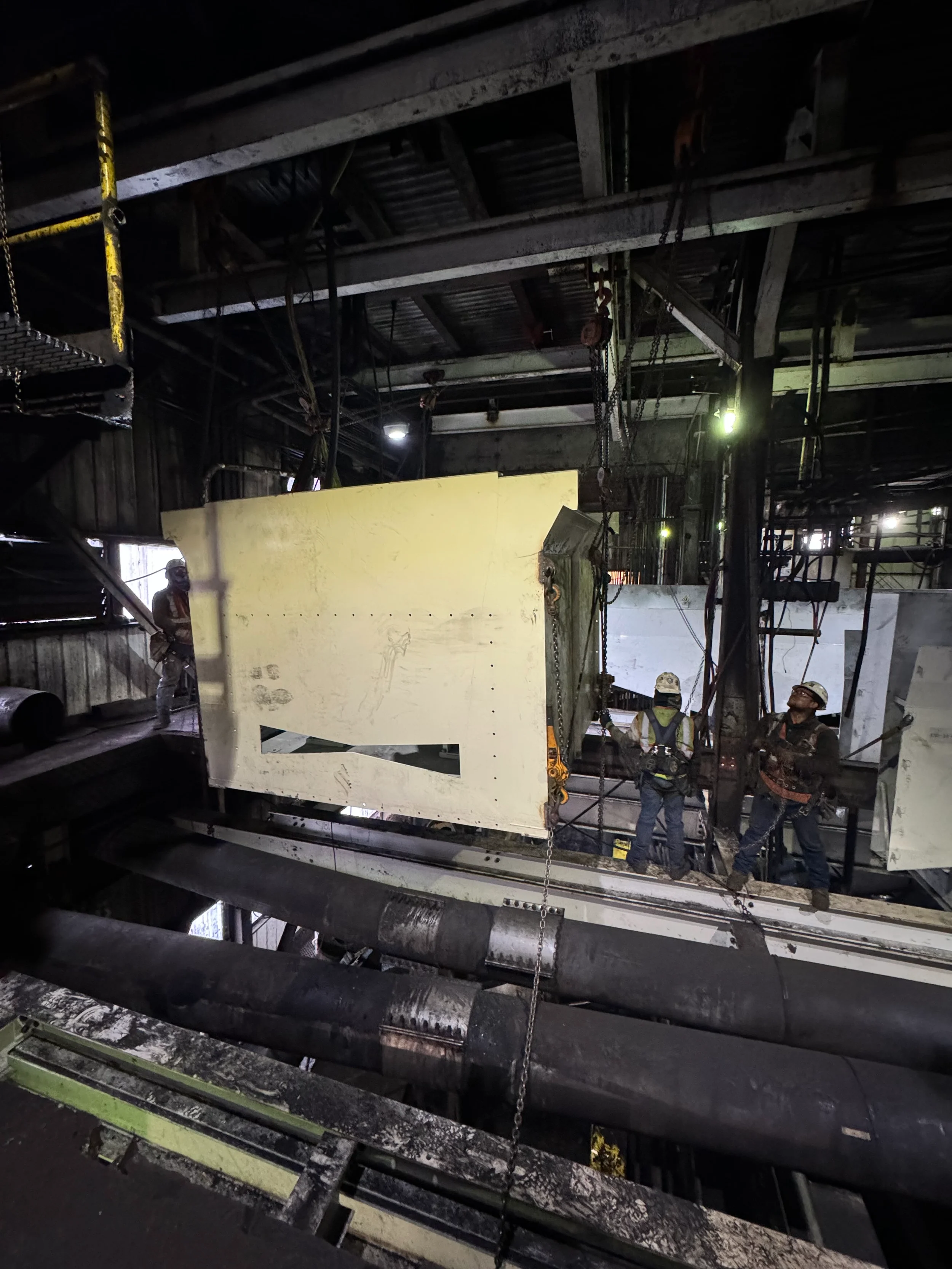 Workers wearing safety helmets and vests are inside an industrial building, working on large metal pipes and equipment in a dimly lit space.