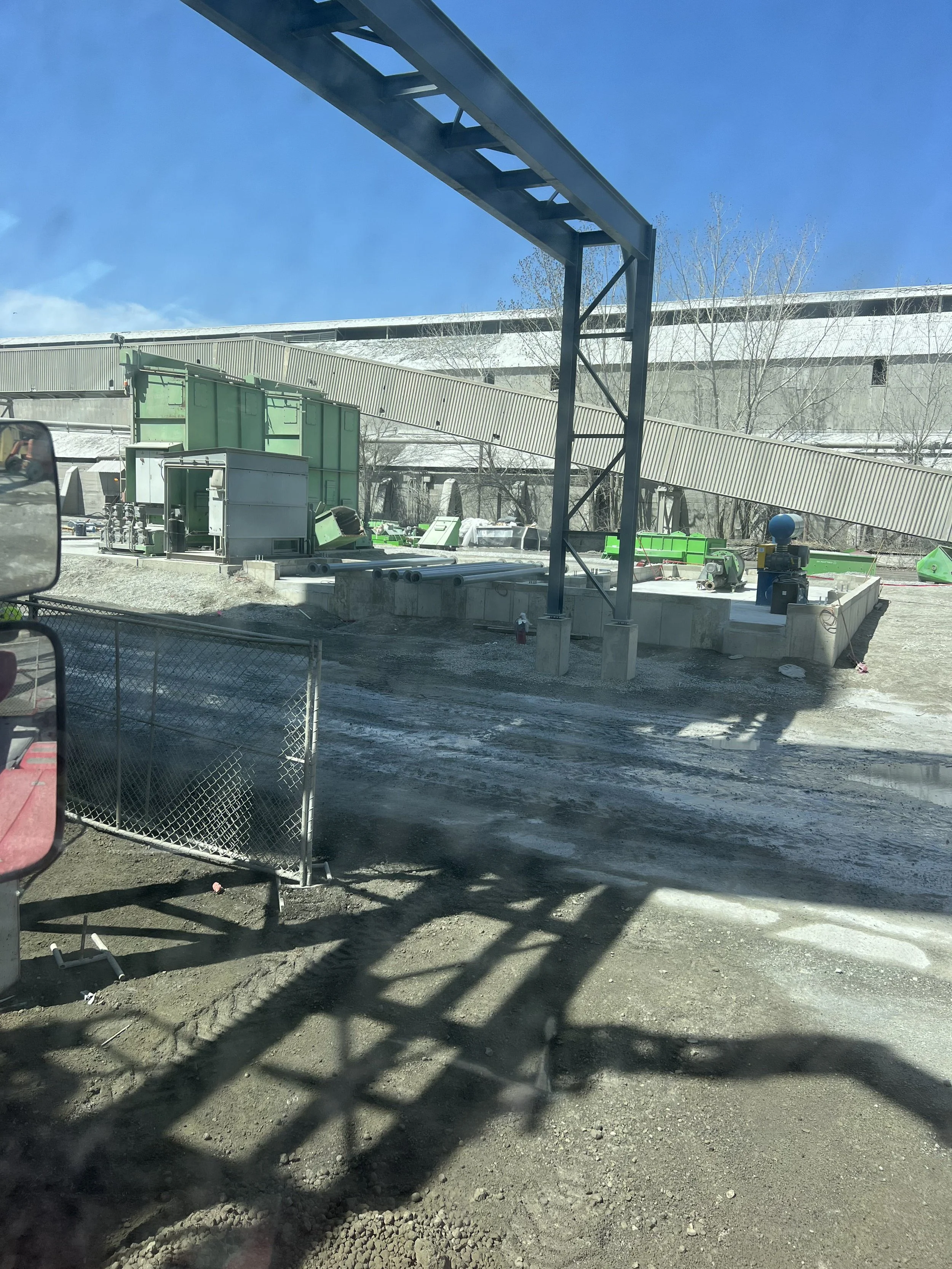 Construction site with equipment, a conveyor belt, and snow-covered ground, viewed through a vehicle window with shadows on the dirt.