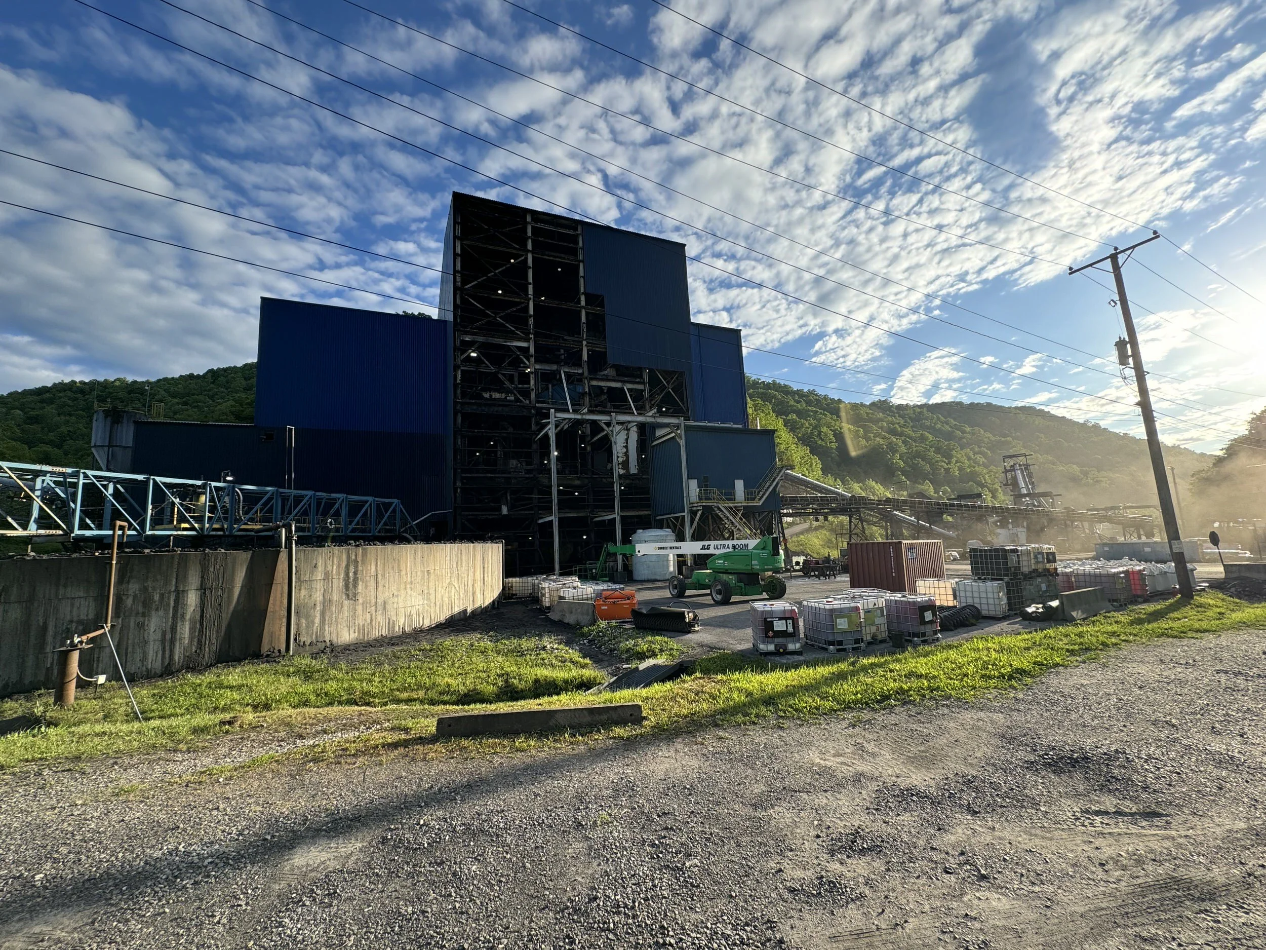 A construction site with large blue metal building structures, construction equipment, and materials under a partly cloudy sky surrounded by green hills.