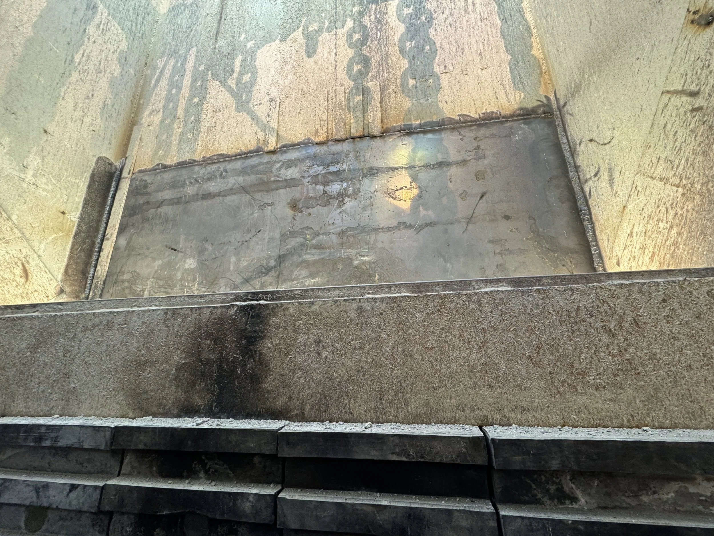 Photo of a metal welding joint on a surface with a chain-like pattern on the wall in the background.