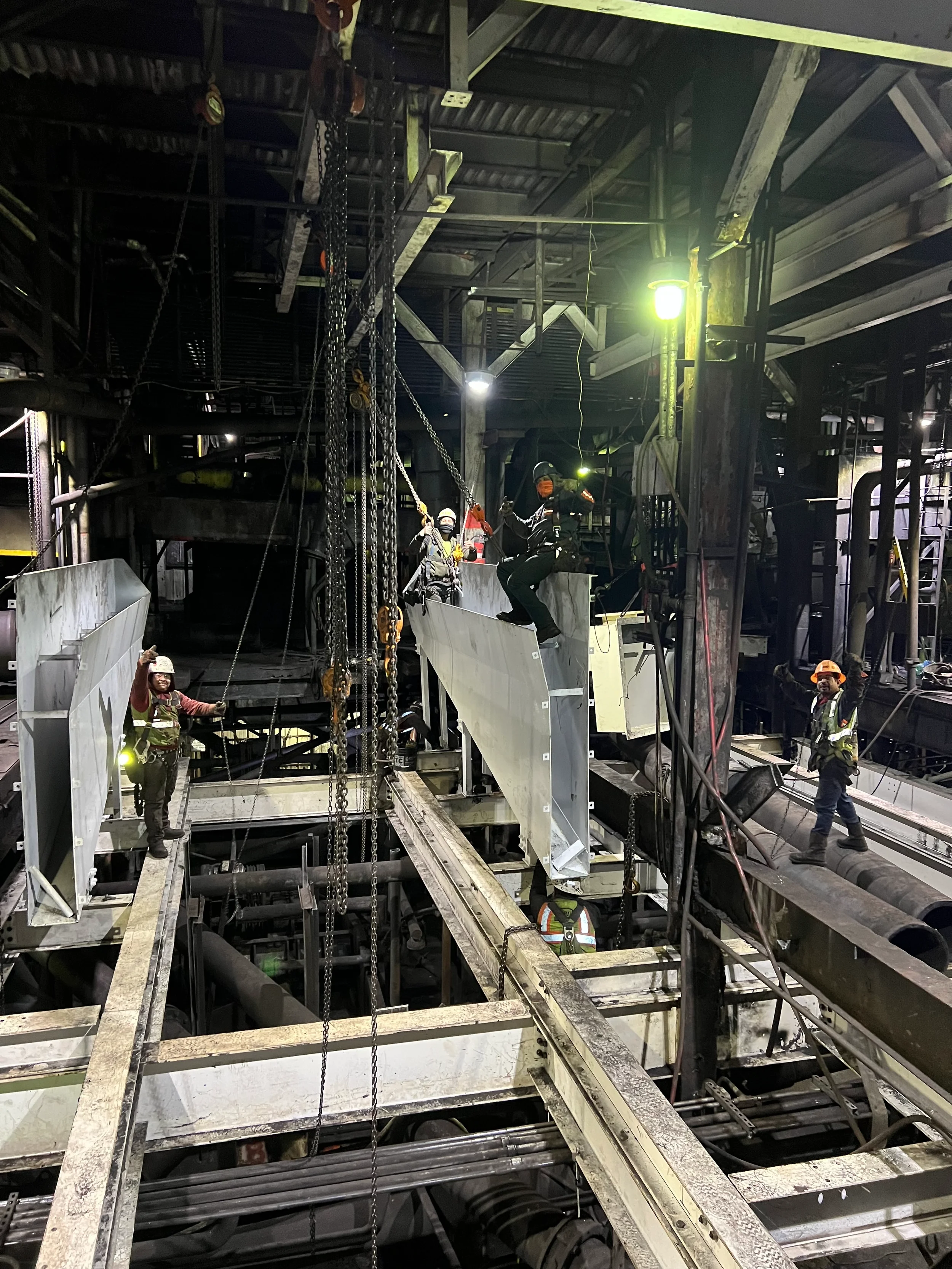 Workers wearing safety gear and helmets working inside an industrial facility with metal walkways, chains, and machinery.