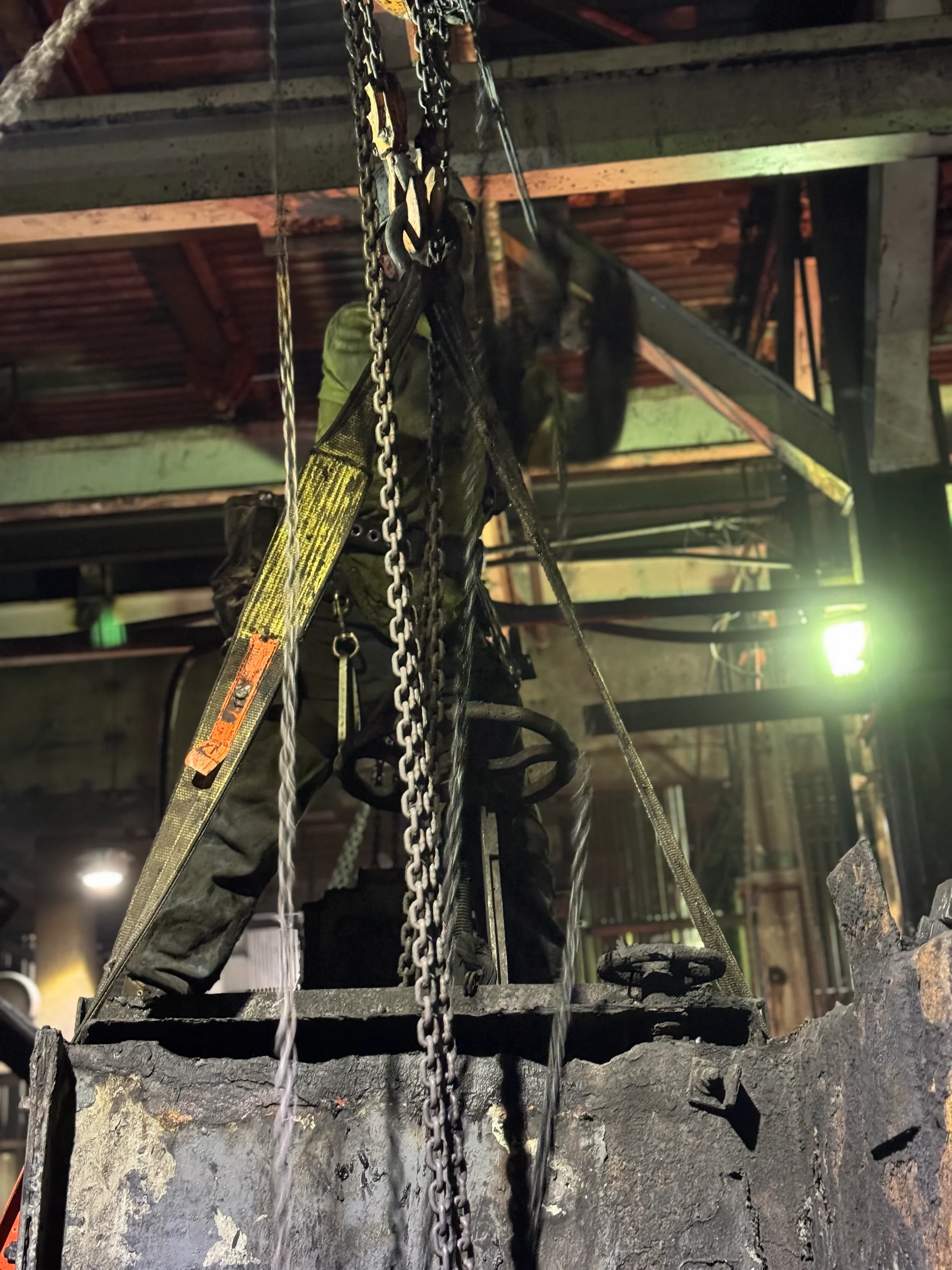 A worker dressed in dark protective gear and gloves on a ladder, hanging chains, and working near a blackened, charred fire pit in an industrial setting with metal beams and concrete walls.
