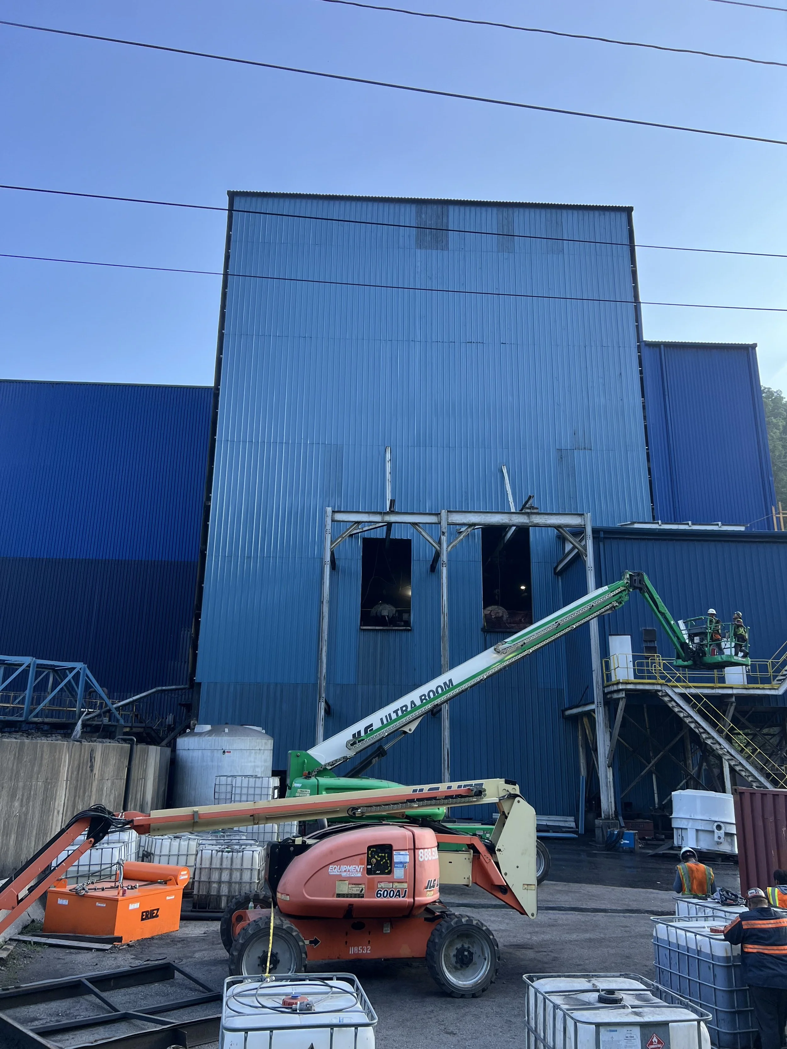 Construction site with workers, blue metal building, and machinery including a lift and containers.