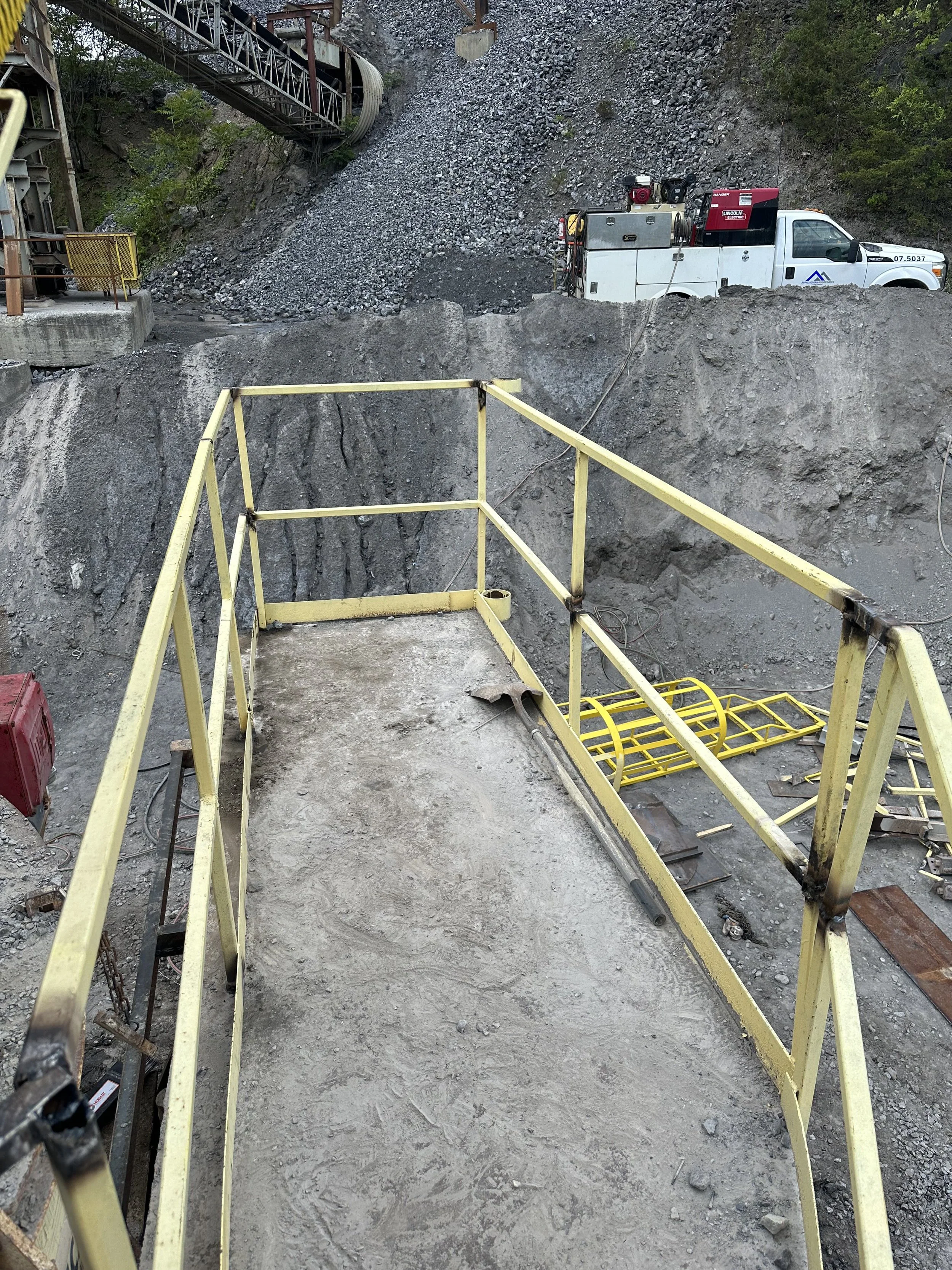 Construction site with a suspended yellow safety railing overlooking a concrete and dirt surface, a work truck with equipment nearby, and a hillside with rubble and a bridge structure in the background.