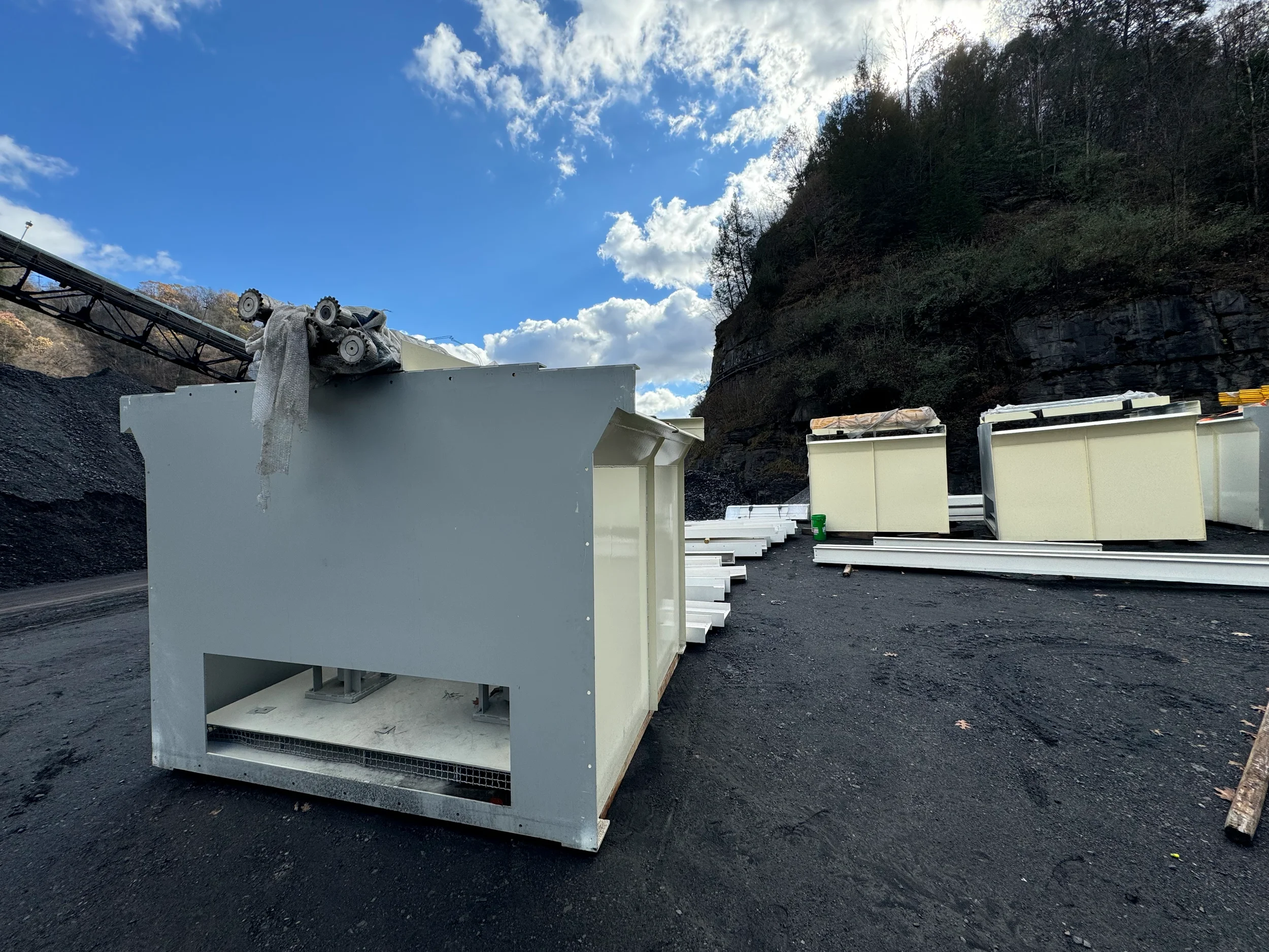Construction materials and equipment outdoors on a dirt surface with a hill covered in trees and a partly cloudy sky in the background.