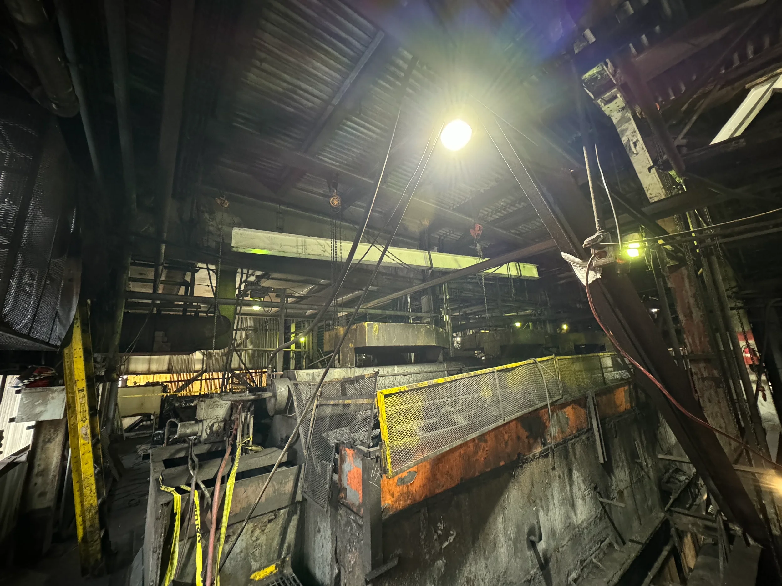 Underground construction site with scaffolding, pipes, and machinery, illuminated by bright lights, with construction barriers and safety tape.