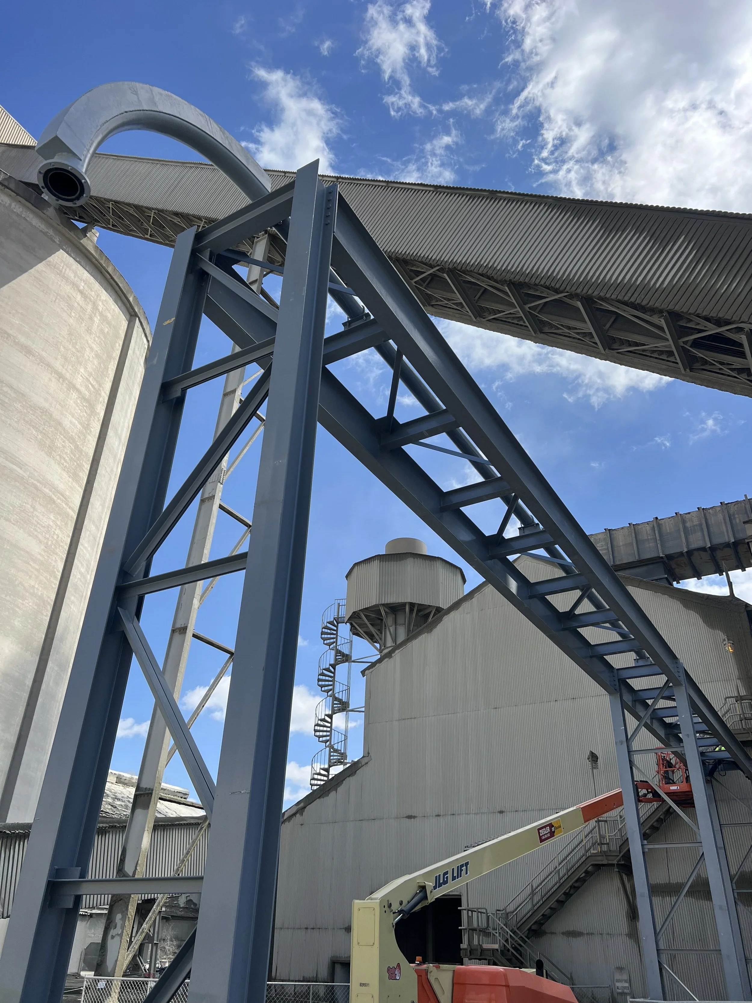 Construction site with large industrial silos, metal framework, and a lift platform, under a blue sky with clouds.