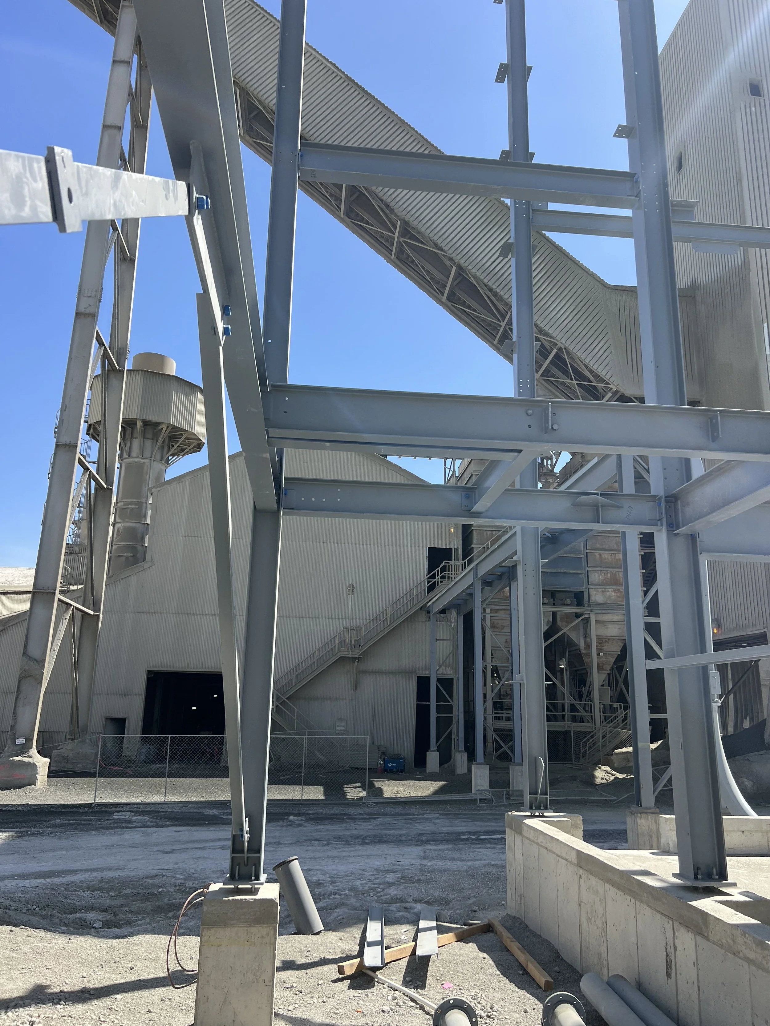Construction site with metal framing, pipes, and stairs, possibly part of an industrial building or factory under construction. Clear blue sky in background.