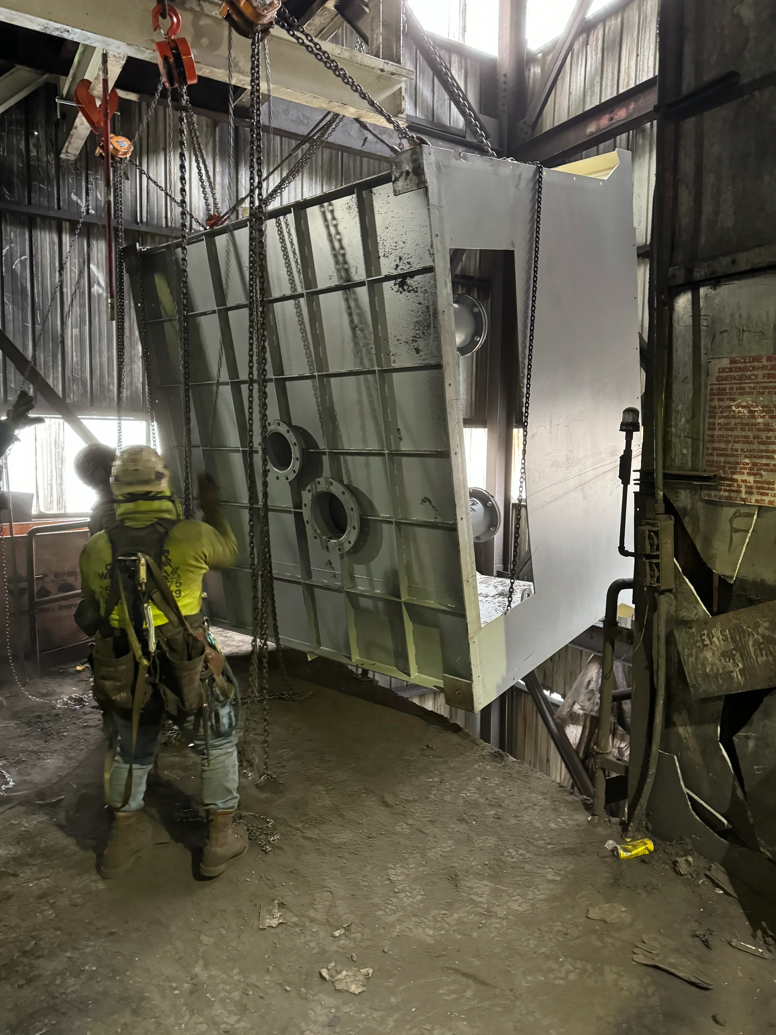 Two construction workers in safety gear working with a large metal structure inside a building with industrial walls and a dirt floor.