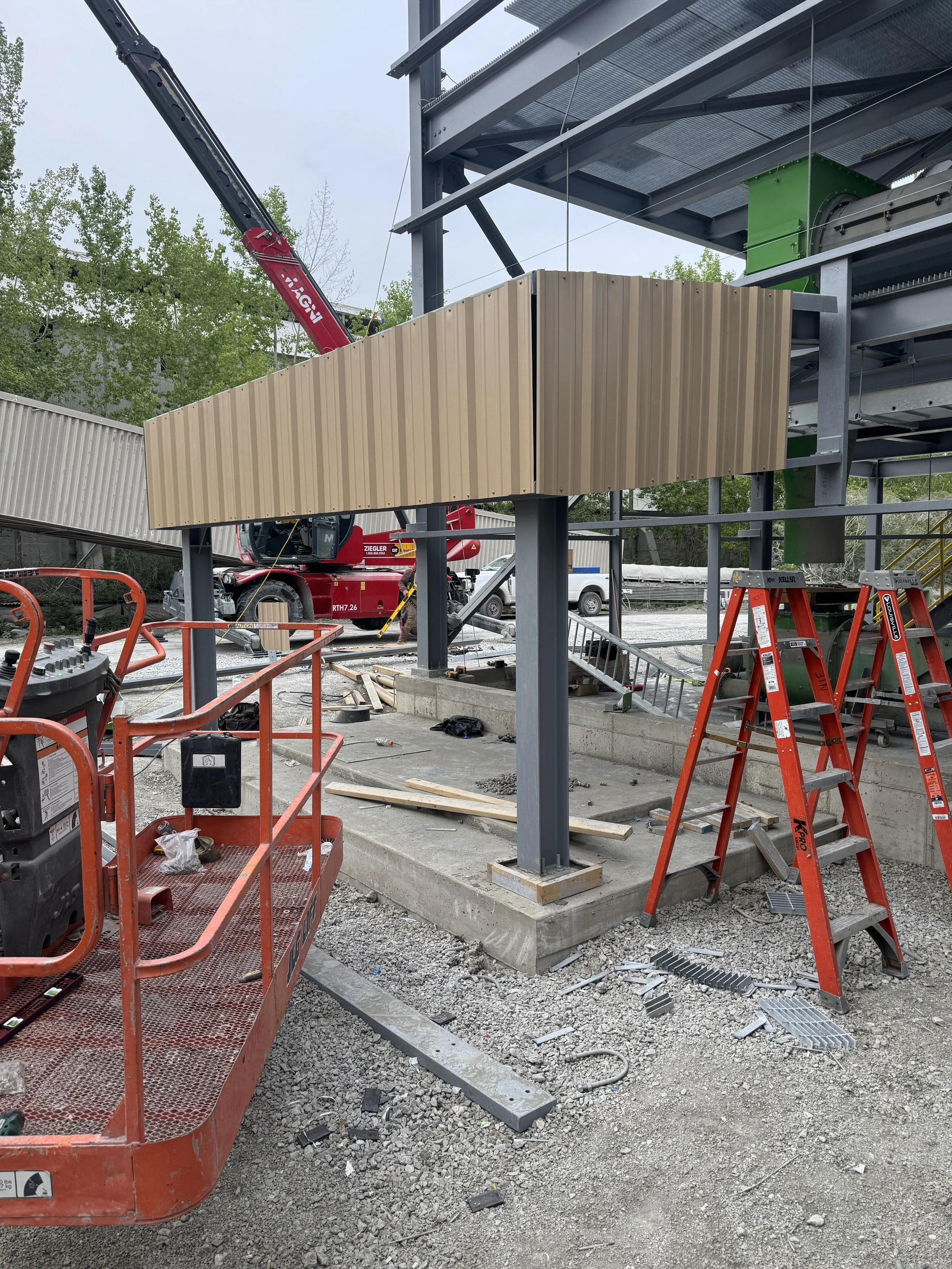 Construction site with steel framework, orange lift, red crane, and corrugated metal panels being installed.