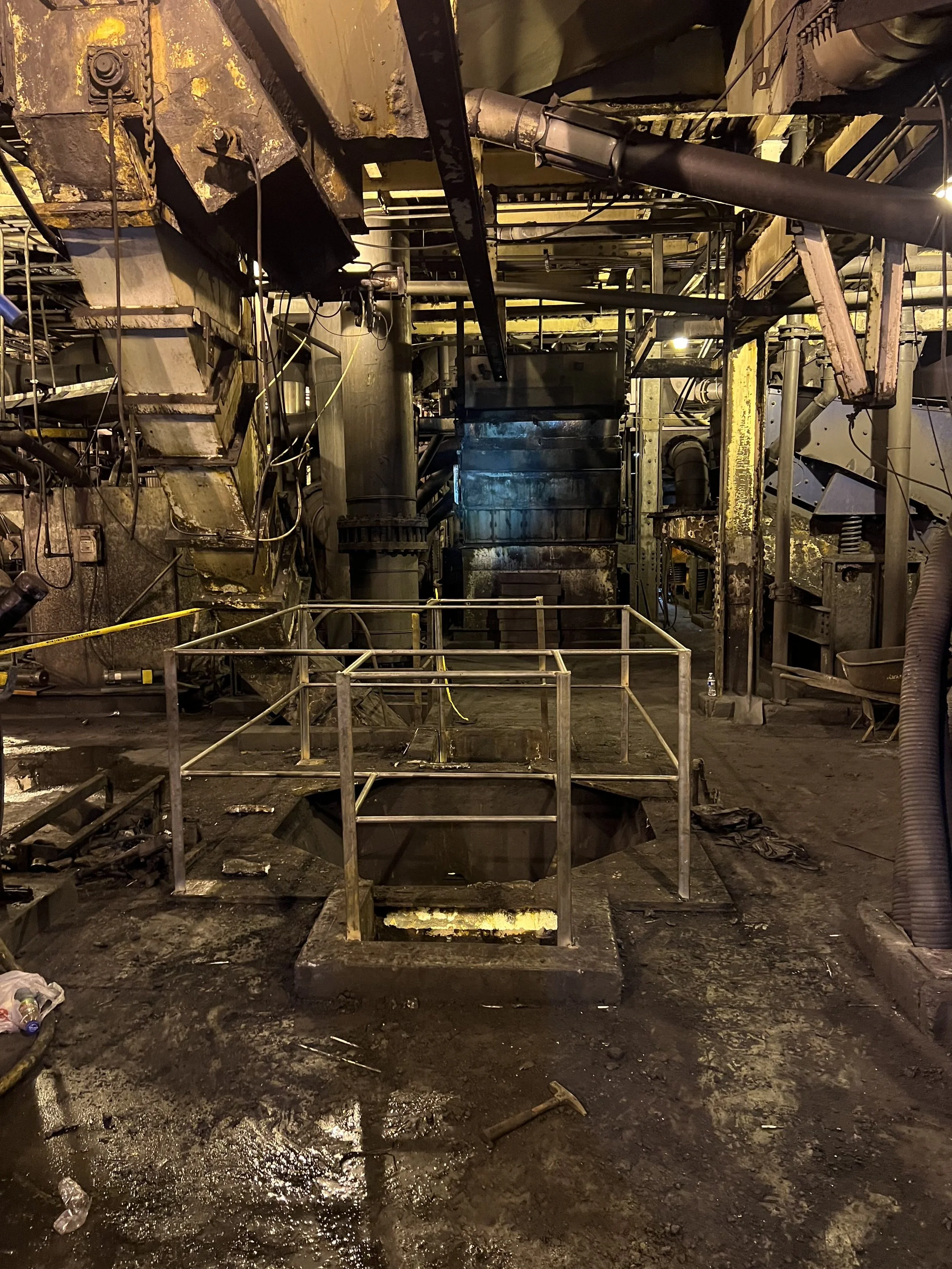 Industrial underground space with metal scaffolding, pipes, and machinery. The floor is muddy, and there are construction barriers and scattered debris.