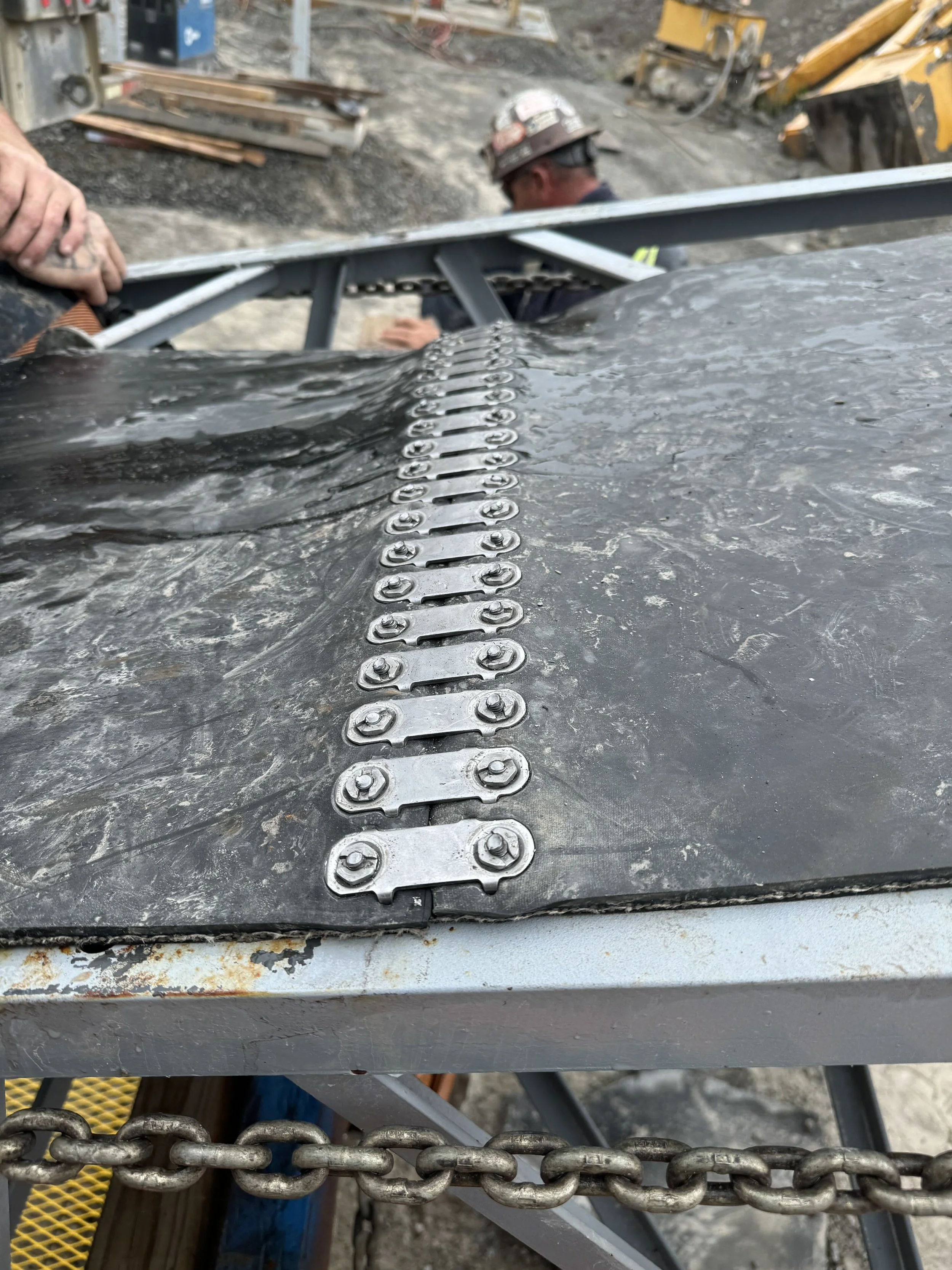 Close-up of a metal structure with chained links and bolts, with construction workers and equipment in the background at a construction site.