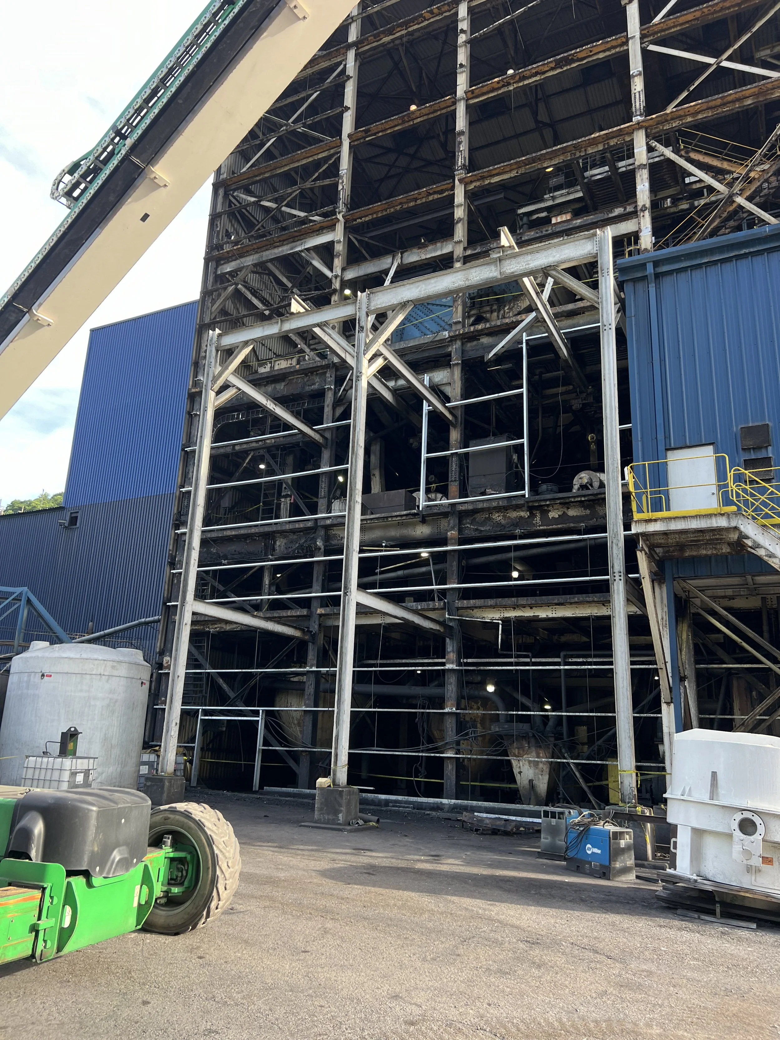 Construction site with a large building framework, scaffolding, and equipment including a green pallet jack, a white tank, and tools on the ground.