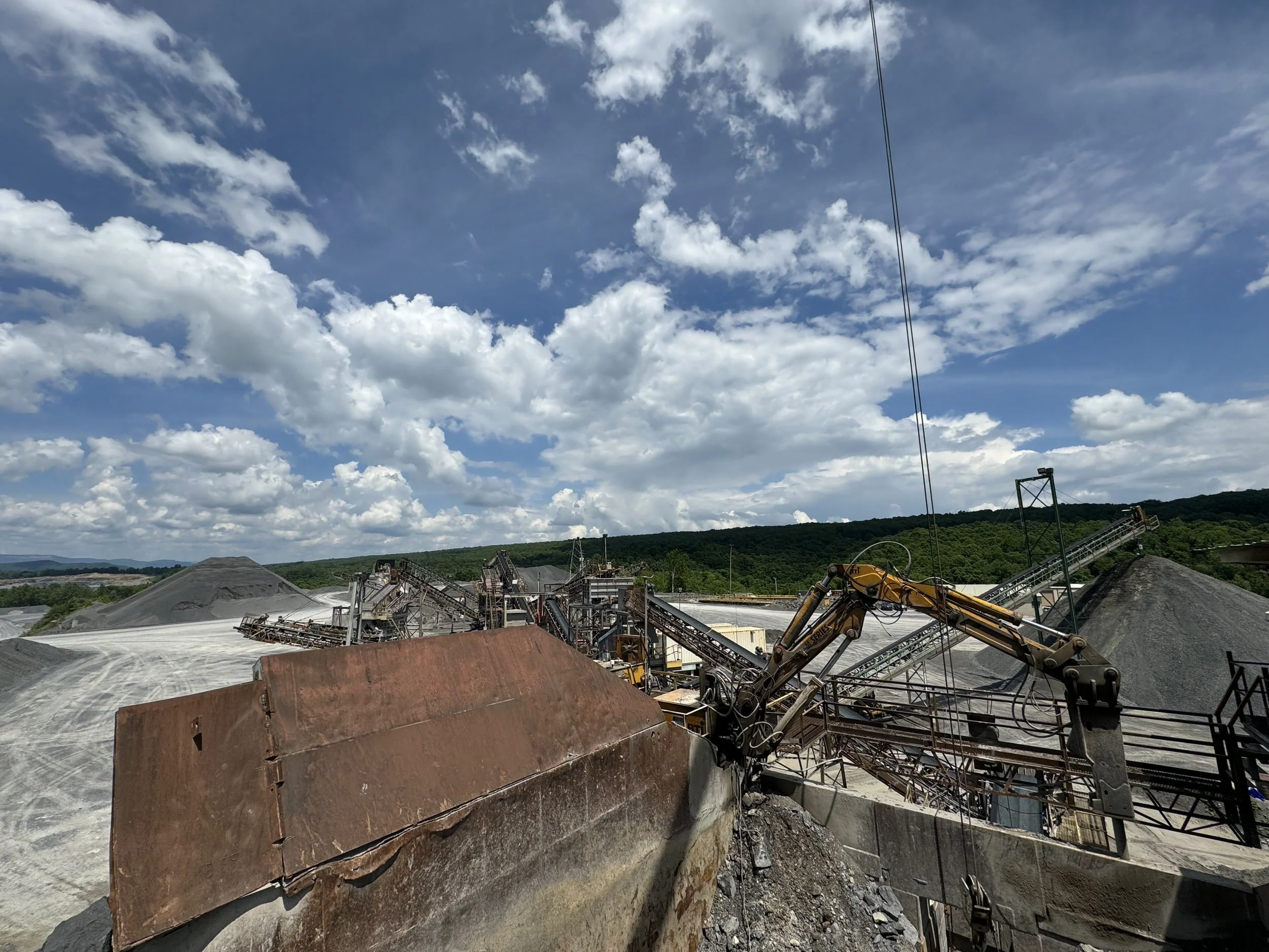 View of an industrial mining or quarry site with large piles of gravel or crushed stone, conveyor belts, and machinery under a partly cloudy sky.