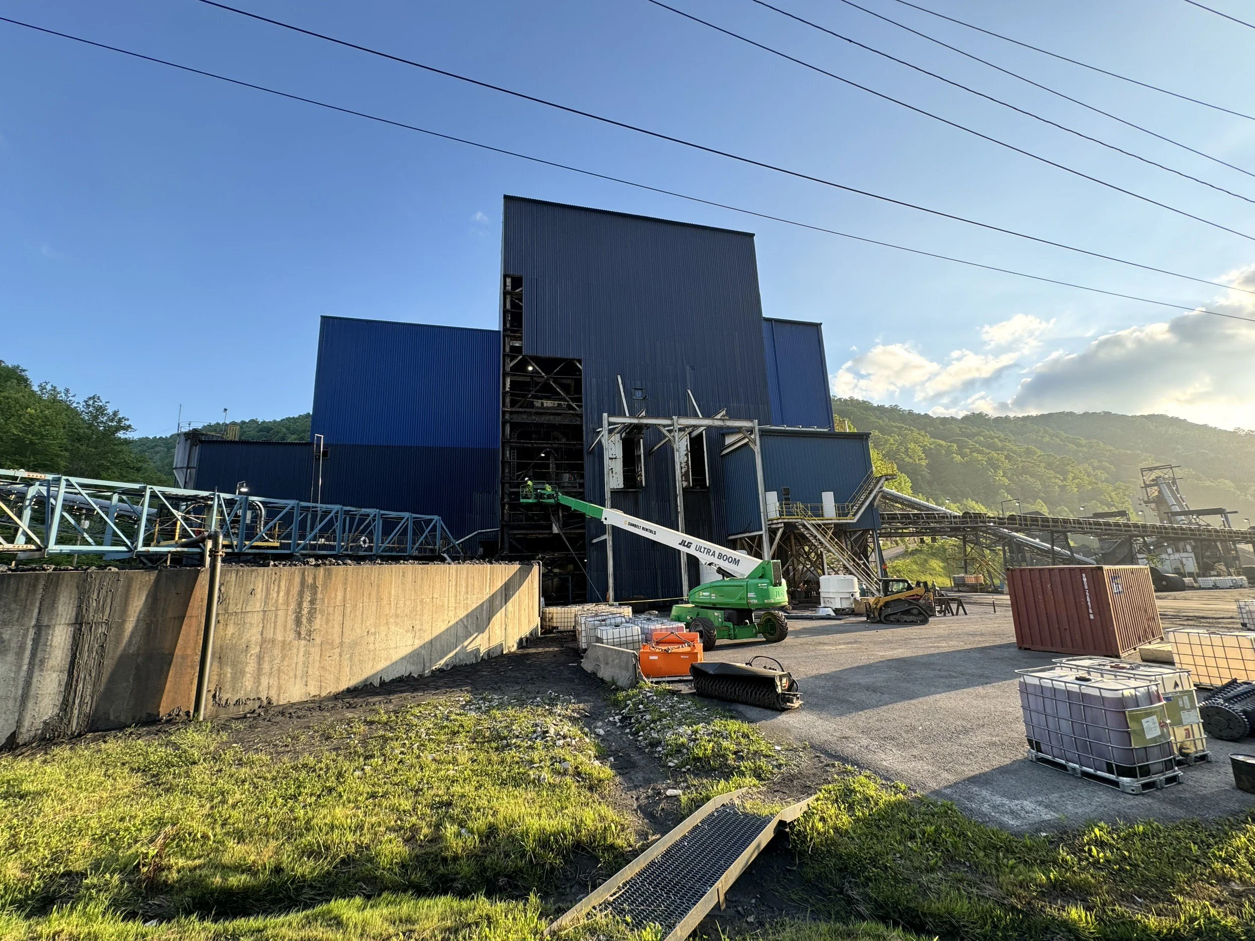 Industrial building with large blue metal walls, construction equipment, and plants in a mountainous area under a partly cloudy sky.