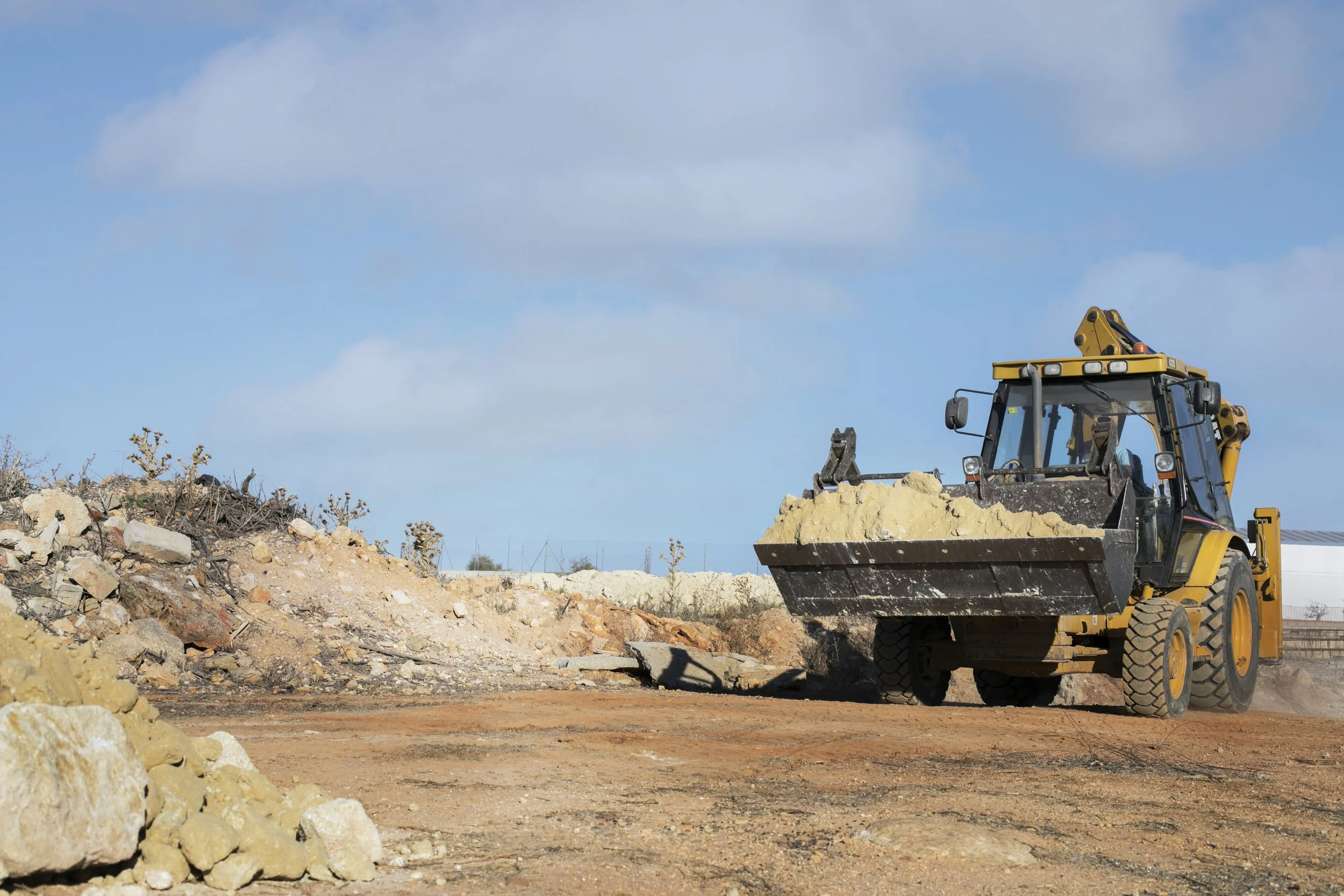 A large yellow construction vehicle, a front loader, is scooping up dirt on a construction site with a partly cloudy sky in the background.