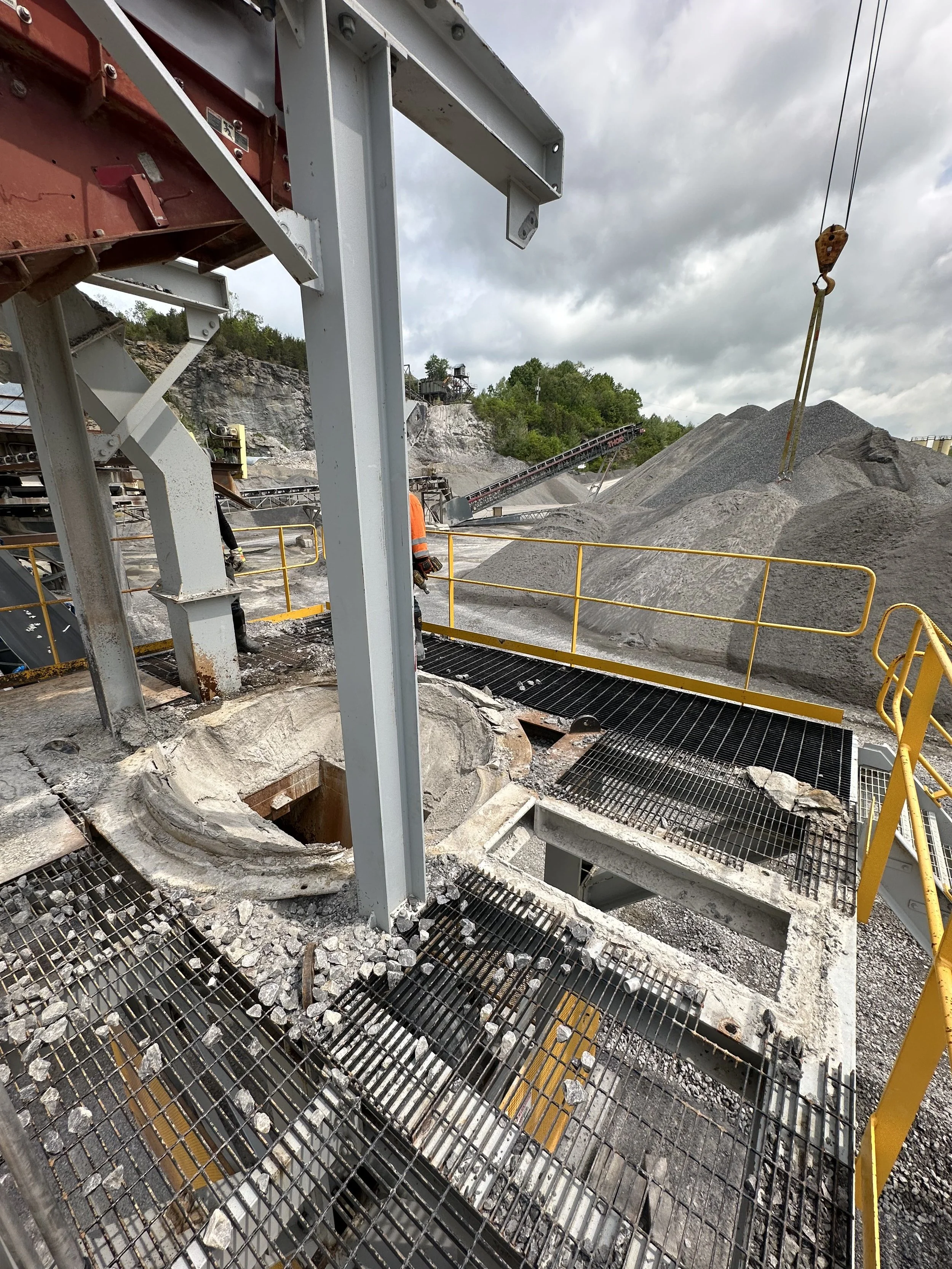 Construction site with steel framework, gravel piles, and workers wearing safety gear under cloudy sky.