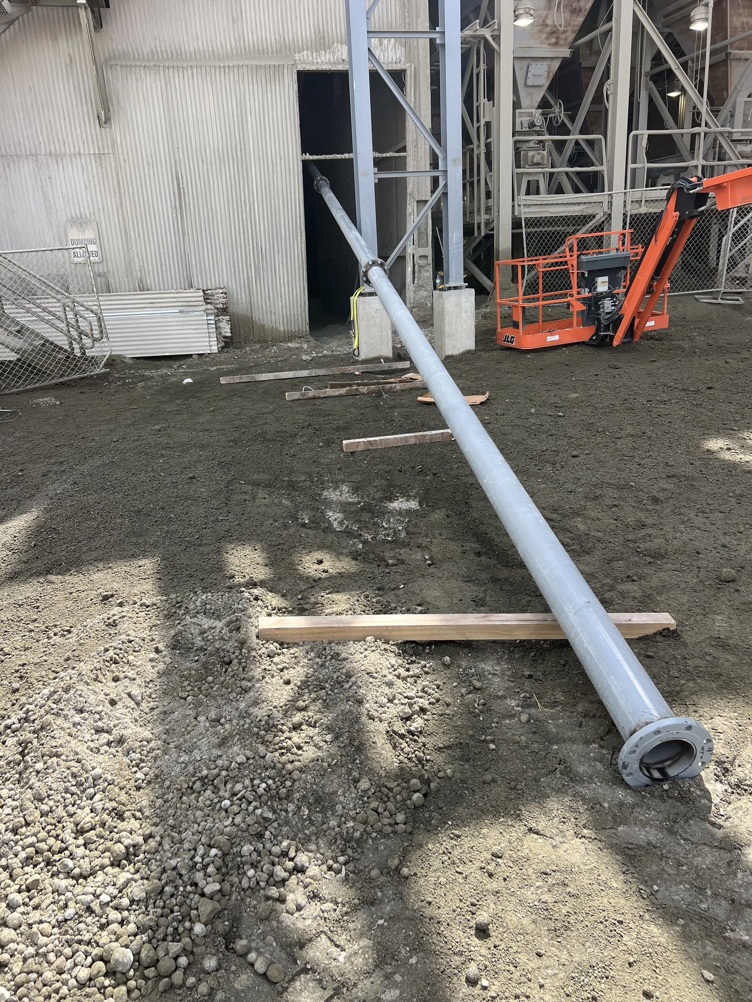 Construction site with a large metal pipe lying on the ground, an orange lift, and a partially constructed building with metal framing and corrugated metal walls.