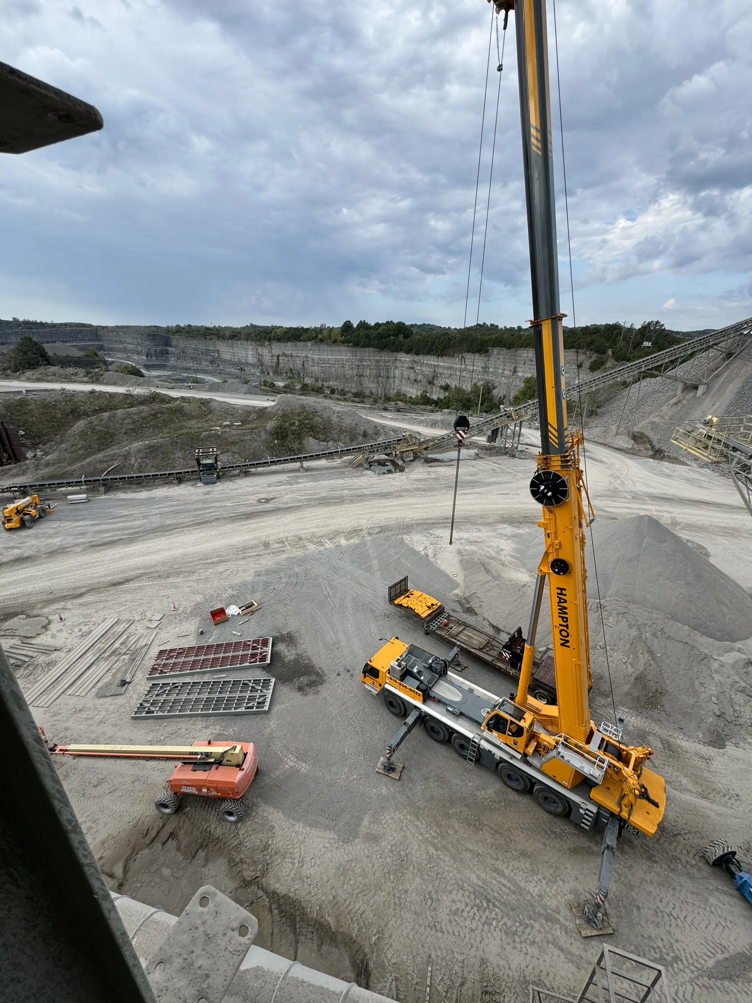 A construction site with a large yellow crane and various construction materials and equipment scattered on the ground, with a quarry or open-pit mine in the background under a cloudy sky.