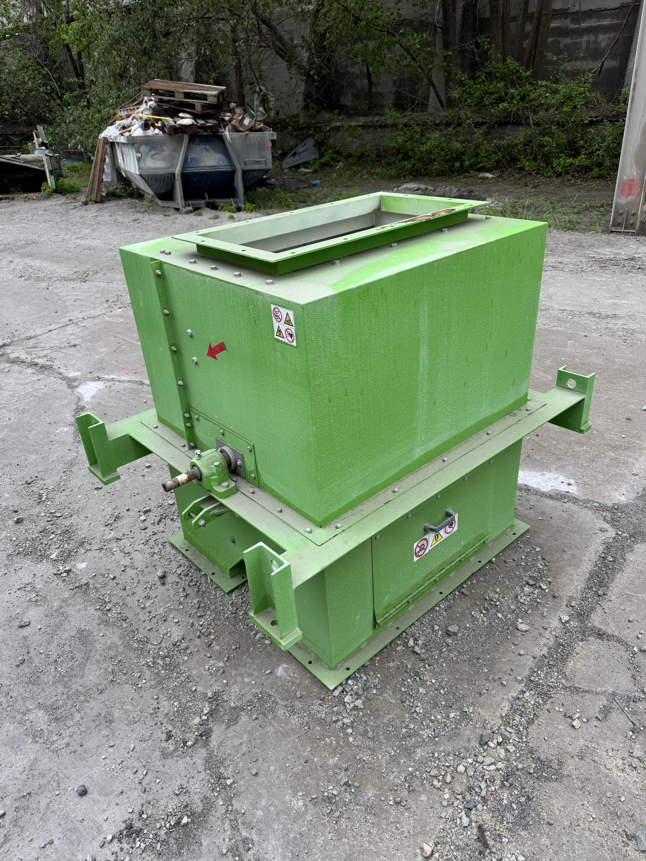 A green industrial shredder machine outdoors on a gravel surface, with warning labels on its side and an open top.