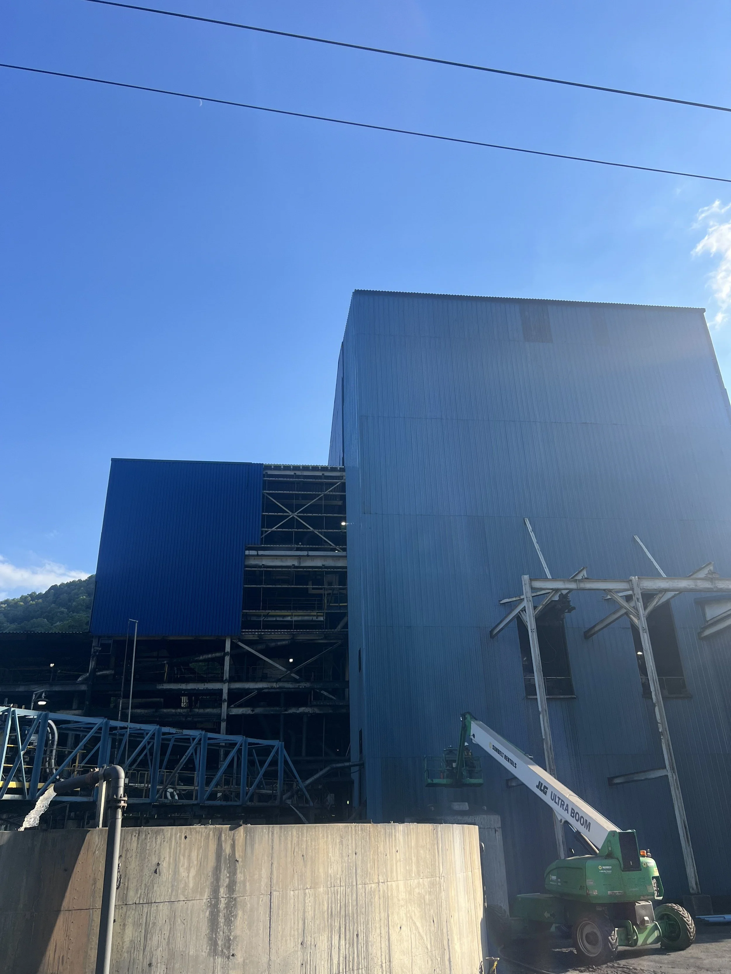 Construction site with blue industrial building, scaffolding, and a cherry picker lift, under a clear blue sky.