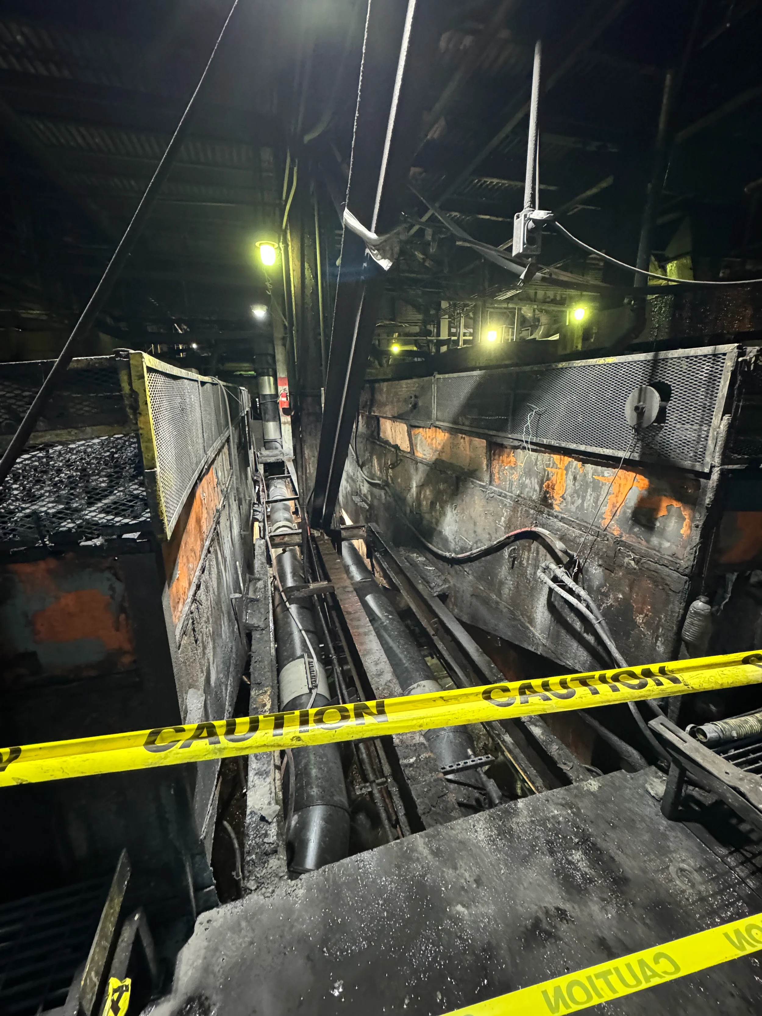 An industrial or construction area with exposed pipes, metal framework, and caution tape. The area looks burned or damaged.