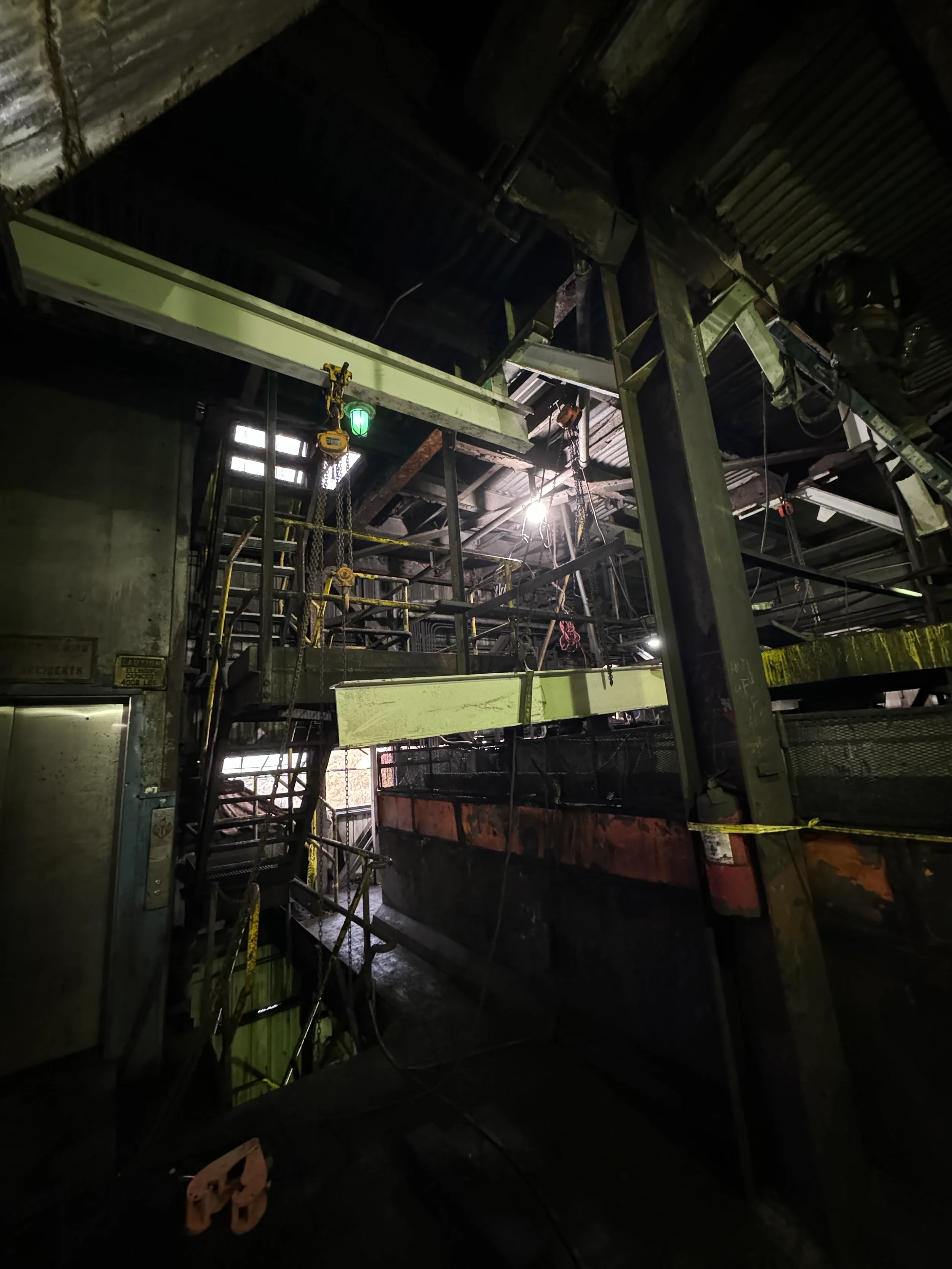 Dark industrial or construction site interior with metal scaffolding, machinery, and yellow caution tape. Some lights and a small green alarm light are visible.