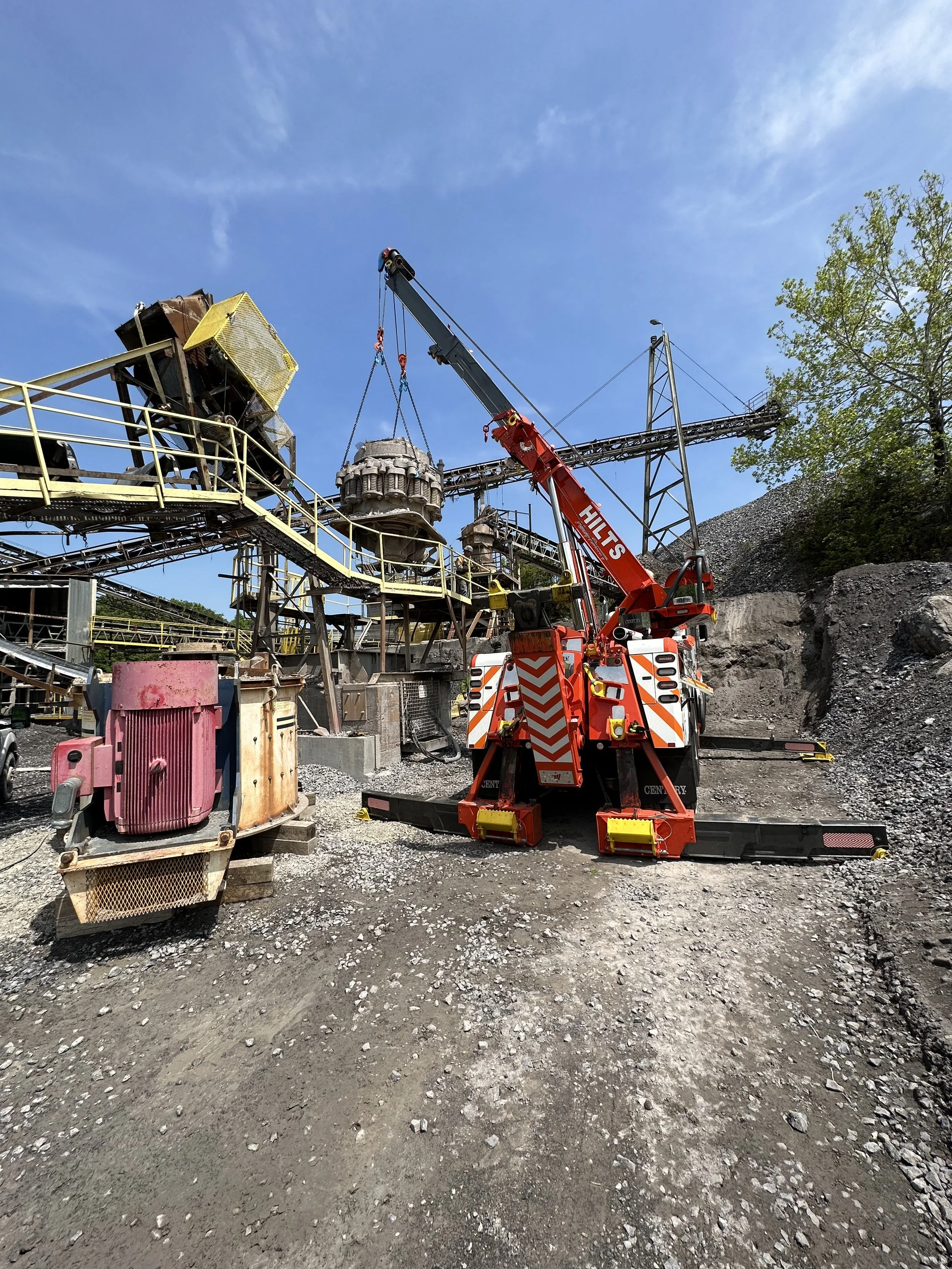 Construction site with a crane lifting a large industrial component, steel structures, and a partly cloudy blue sky.