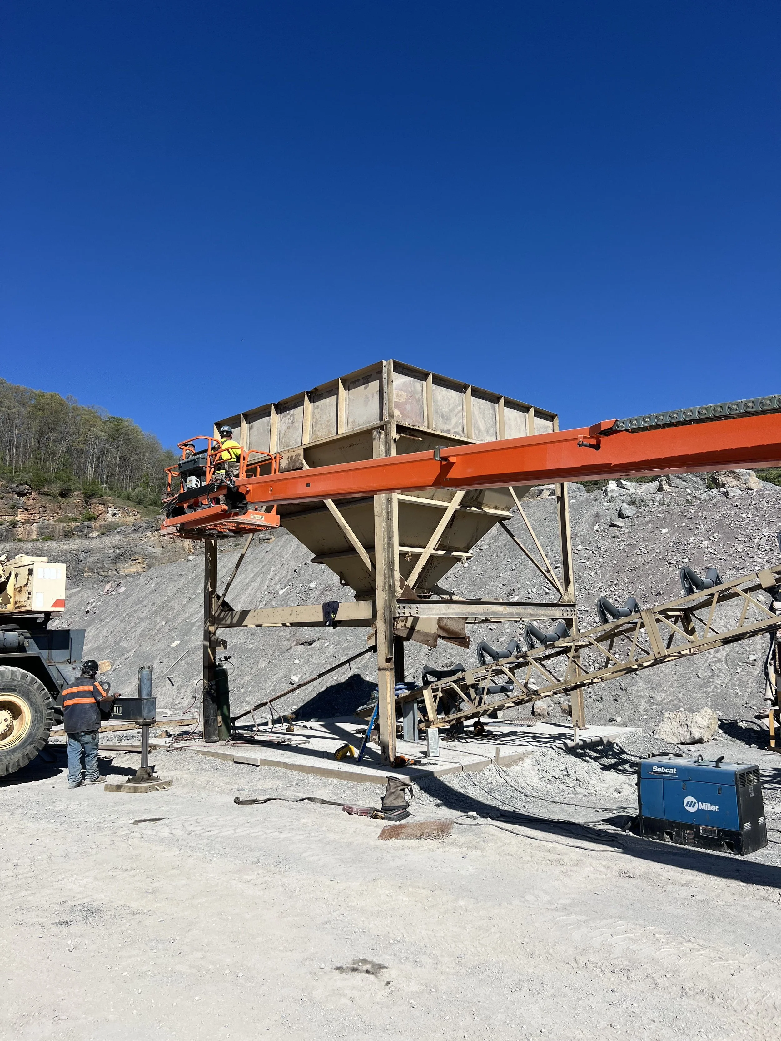 Construction workers operating a cherry picker at a construction site with a large hopper and conveyor system, with rocky terrain and a clear blue sky in the background.