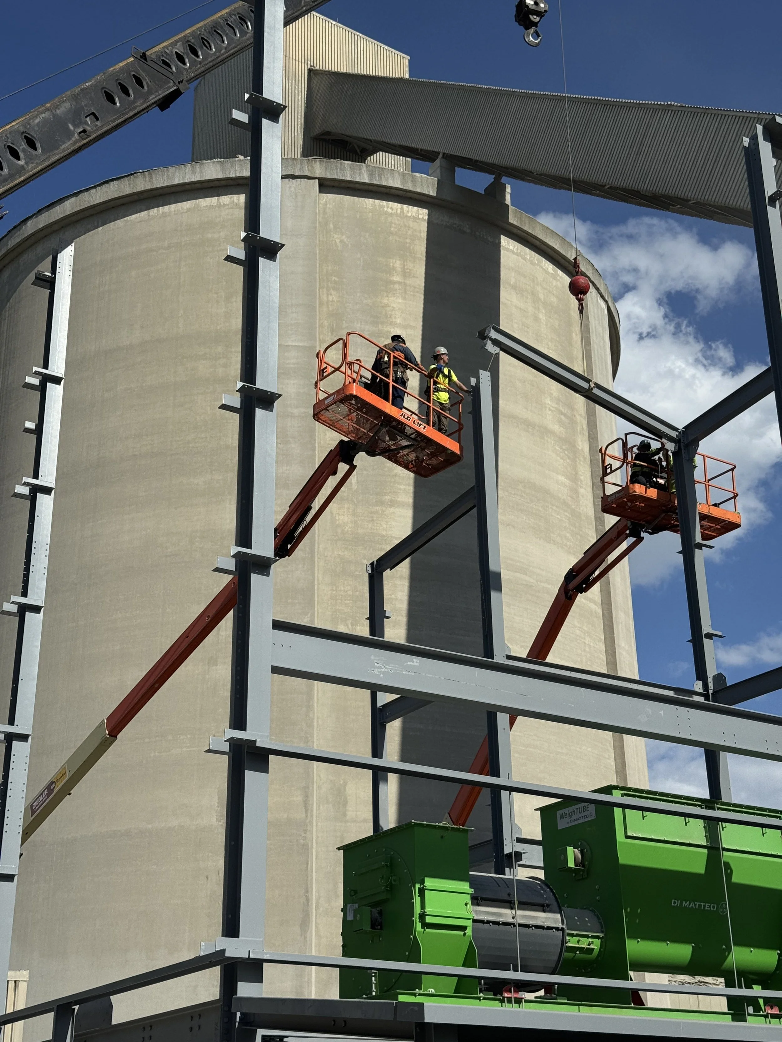 Construction workers on a lift working on a steel framework in front of a large industrial building with a curved concrete wall, blue sky, and some clouds.