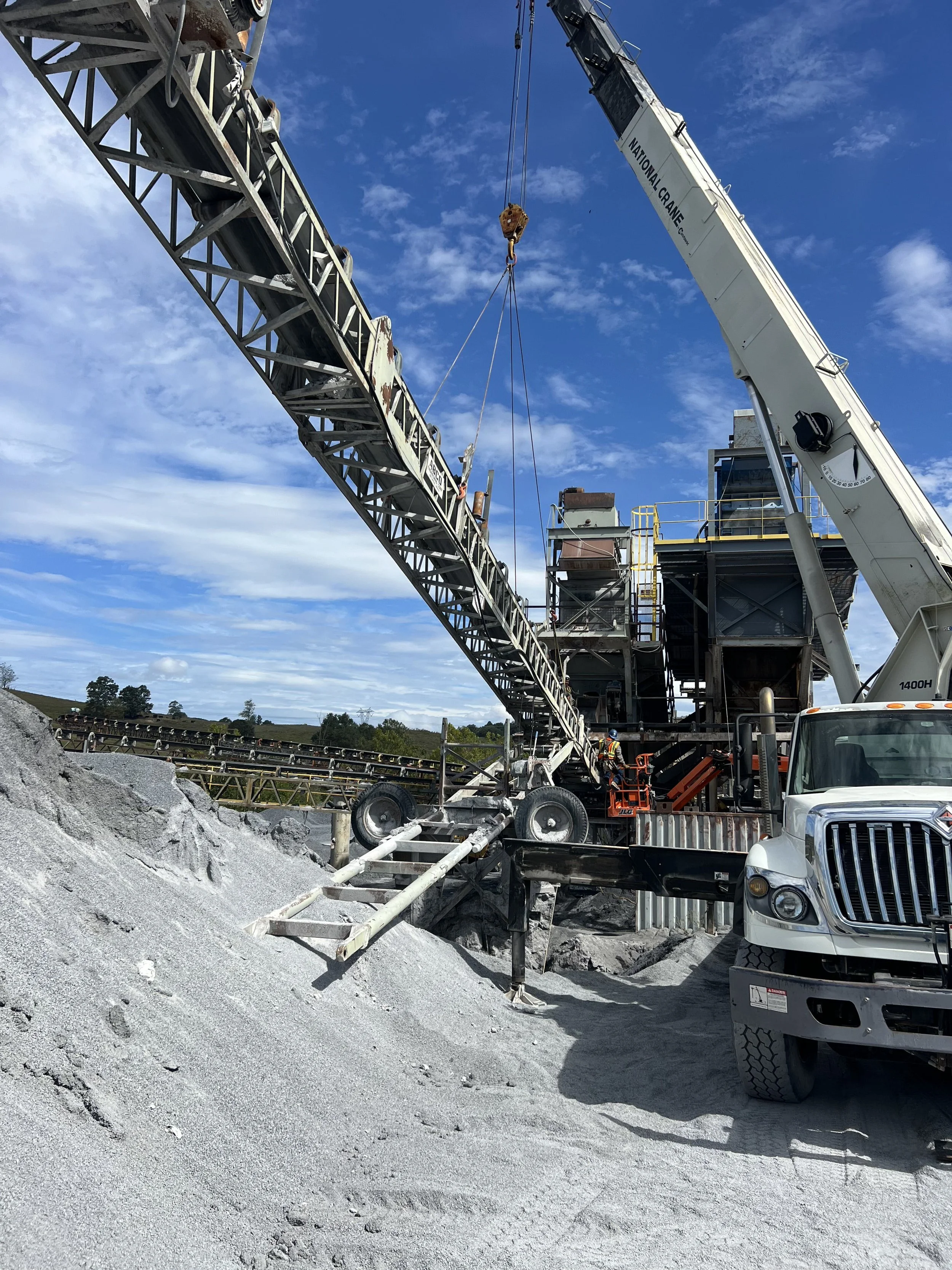 Construction site with a crane lifting materials, a truck, and construction workers working near a rocky area under a blue sky.