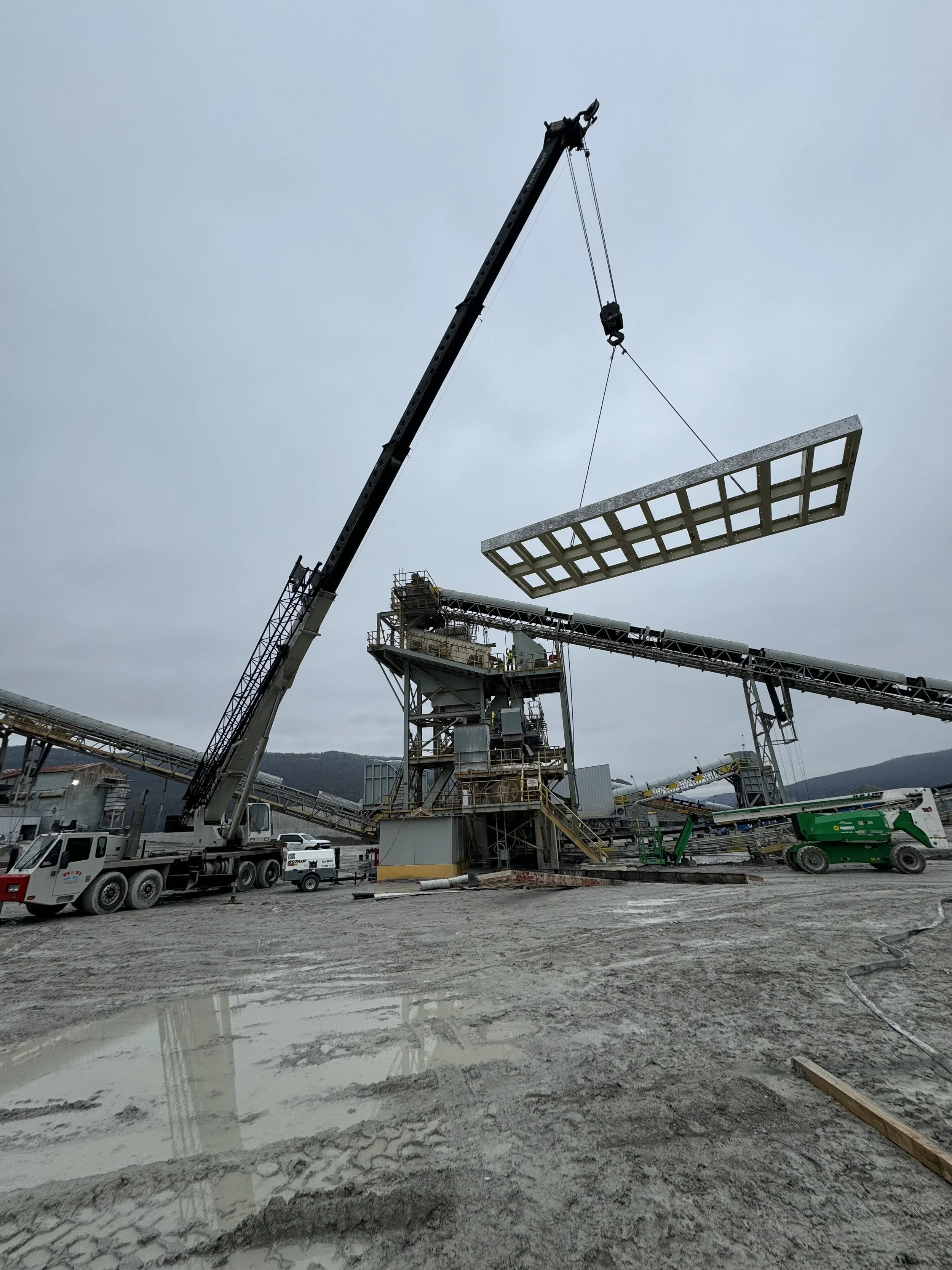 Heavy construction equipment at a construction site, including a crane lifting a large concrete panel, with muddy ground and puddles of water in the foreground.