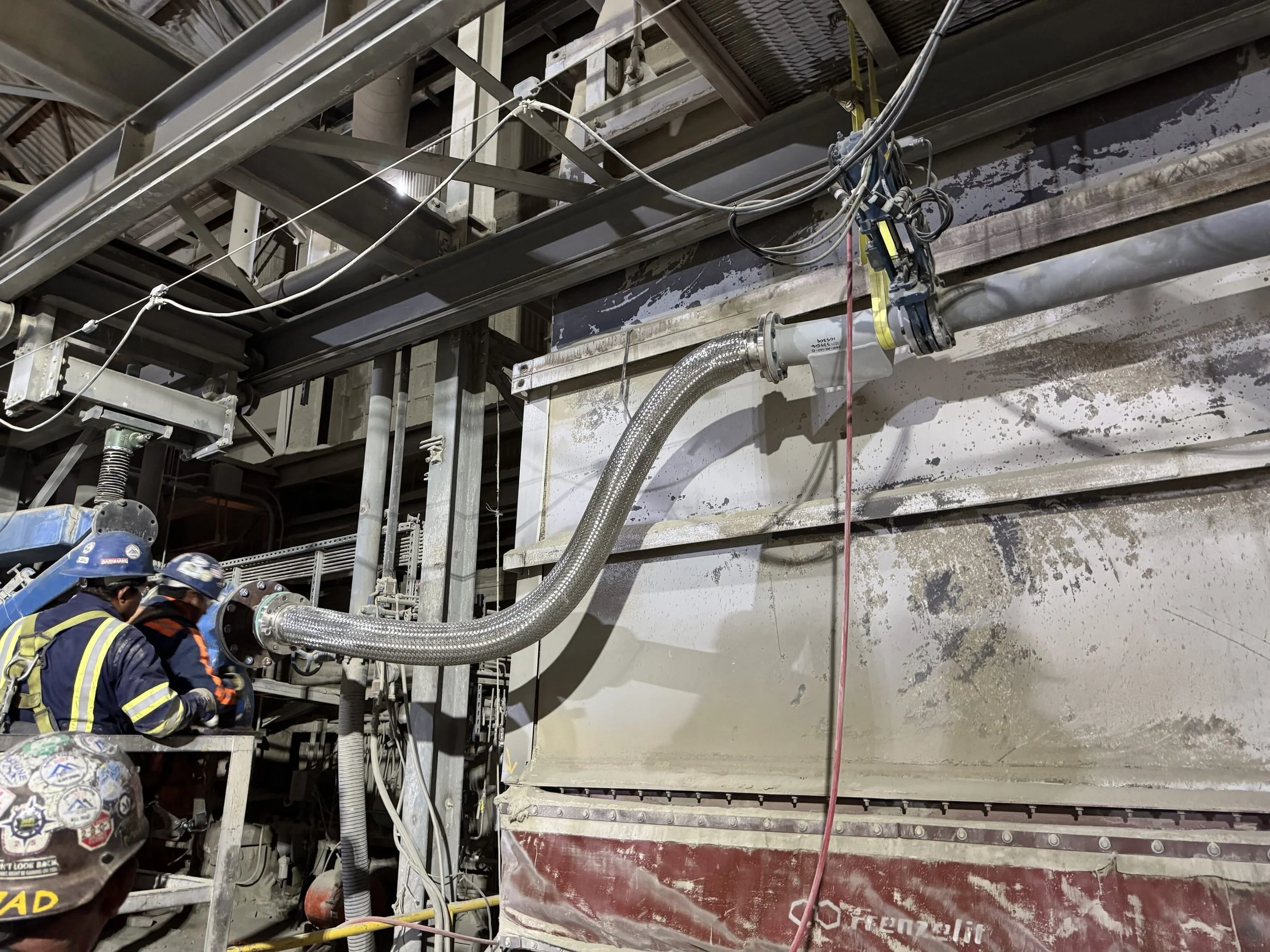 Construction workers in helmets working with pipes and machinery in an industrial setting under scaffolding.