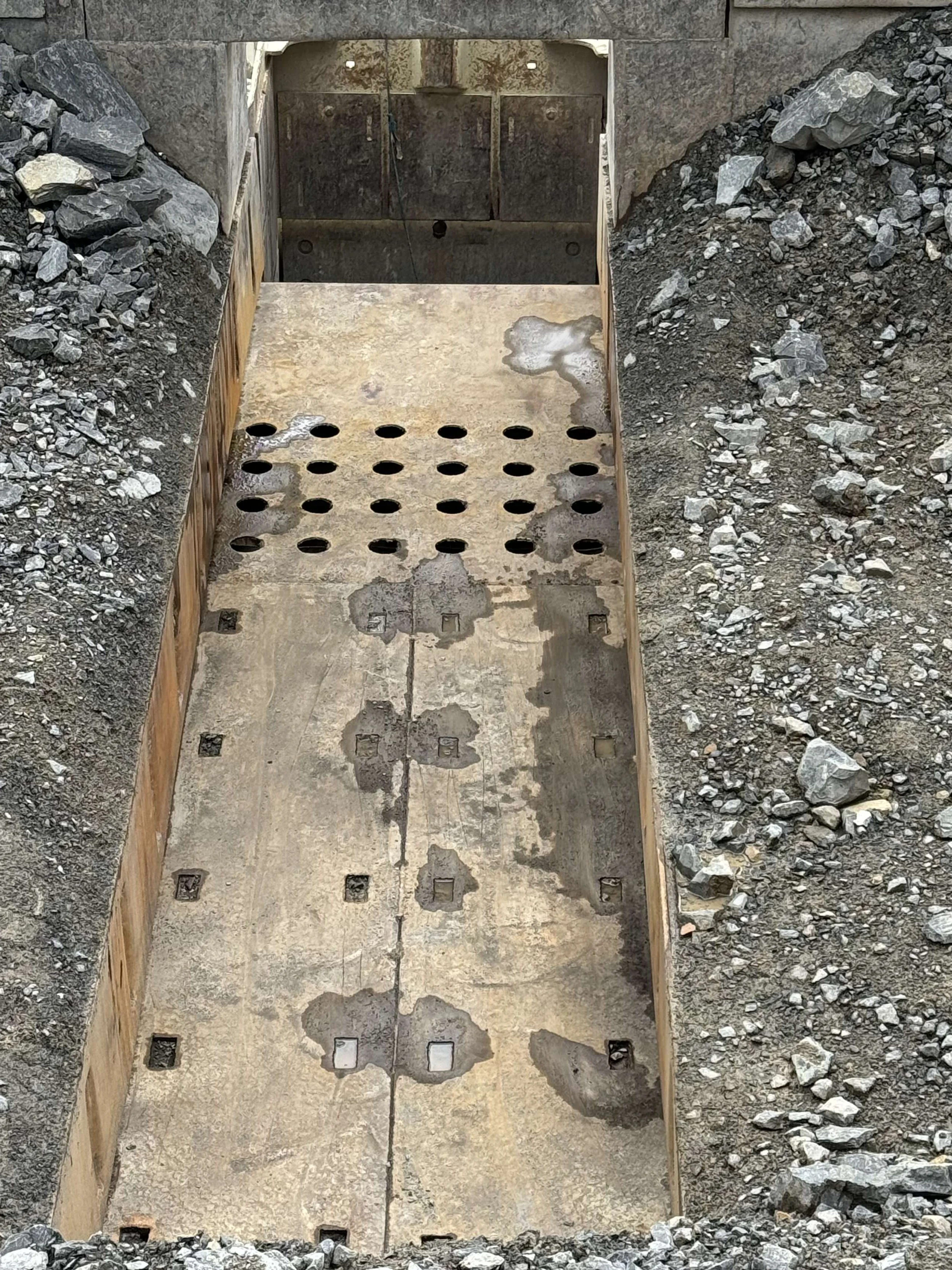 Underground utility or drainage trench with concrete base and holes for pipes, surrounded by soil and rocks.