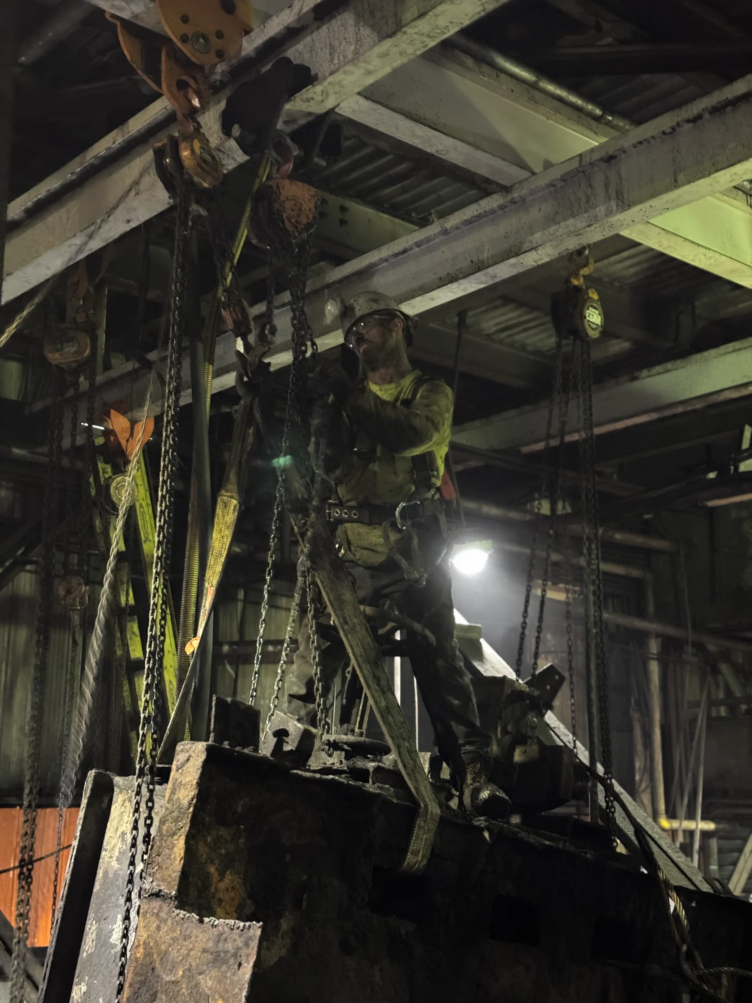 A construction worker wearing safety gear and a helmet standing on a ladder, working amidst chains and metal scaffolding in an industrial setting.