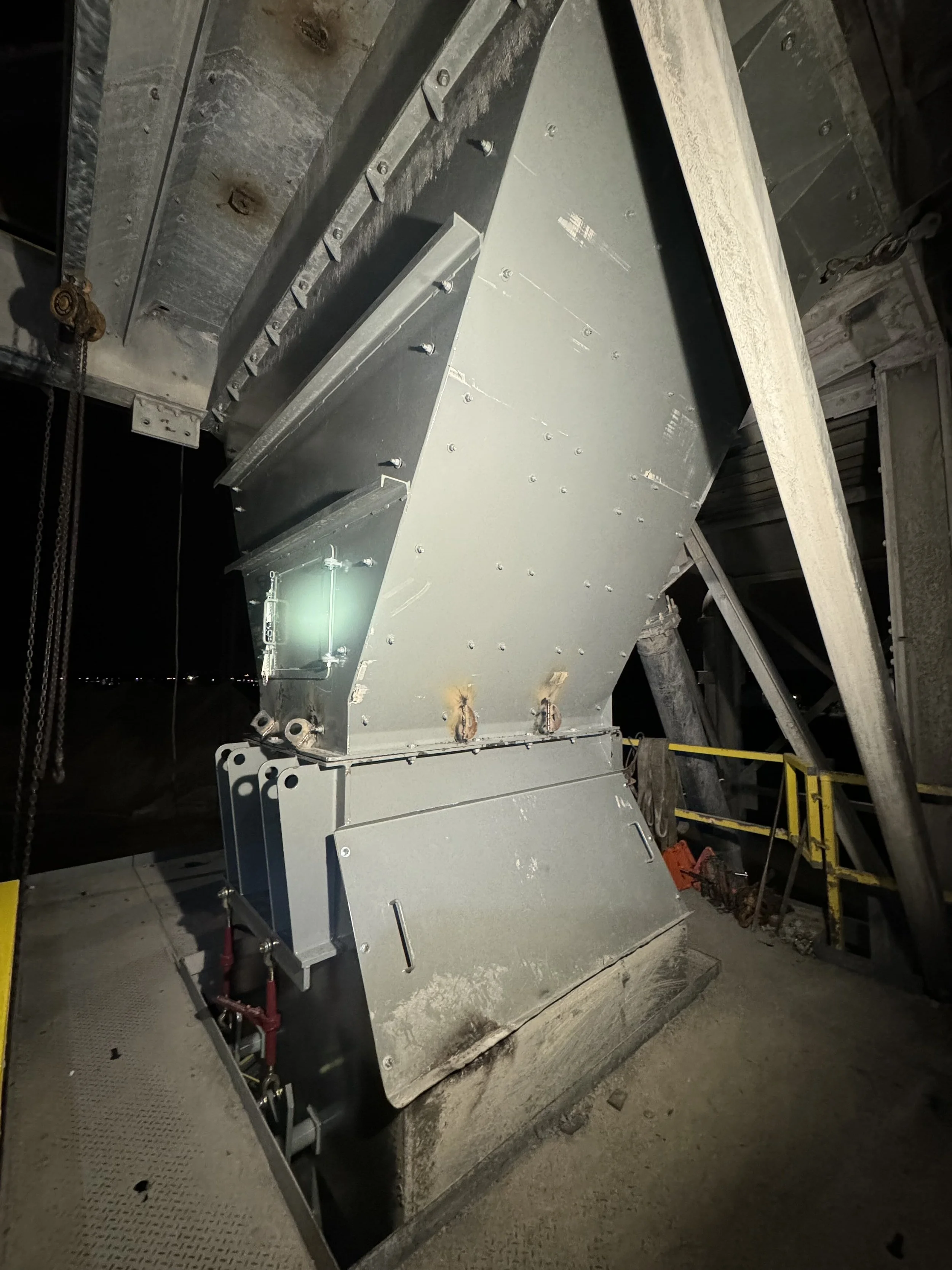 A large metallic industrial machine or component, with various bolts, pipes, and structural elements, located in an industrial or construction setting.