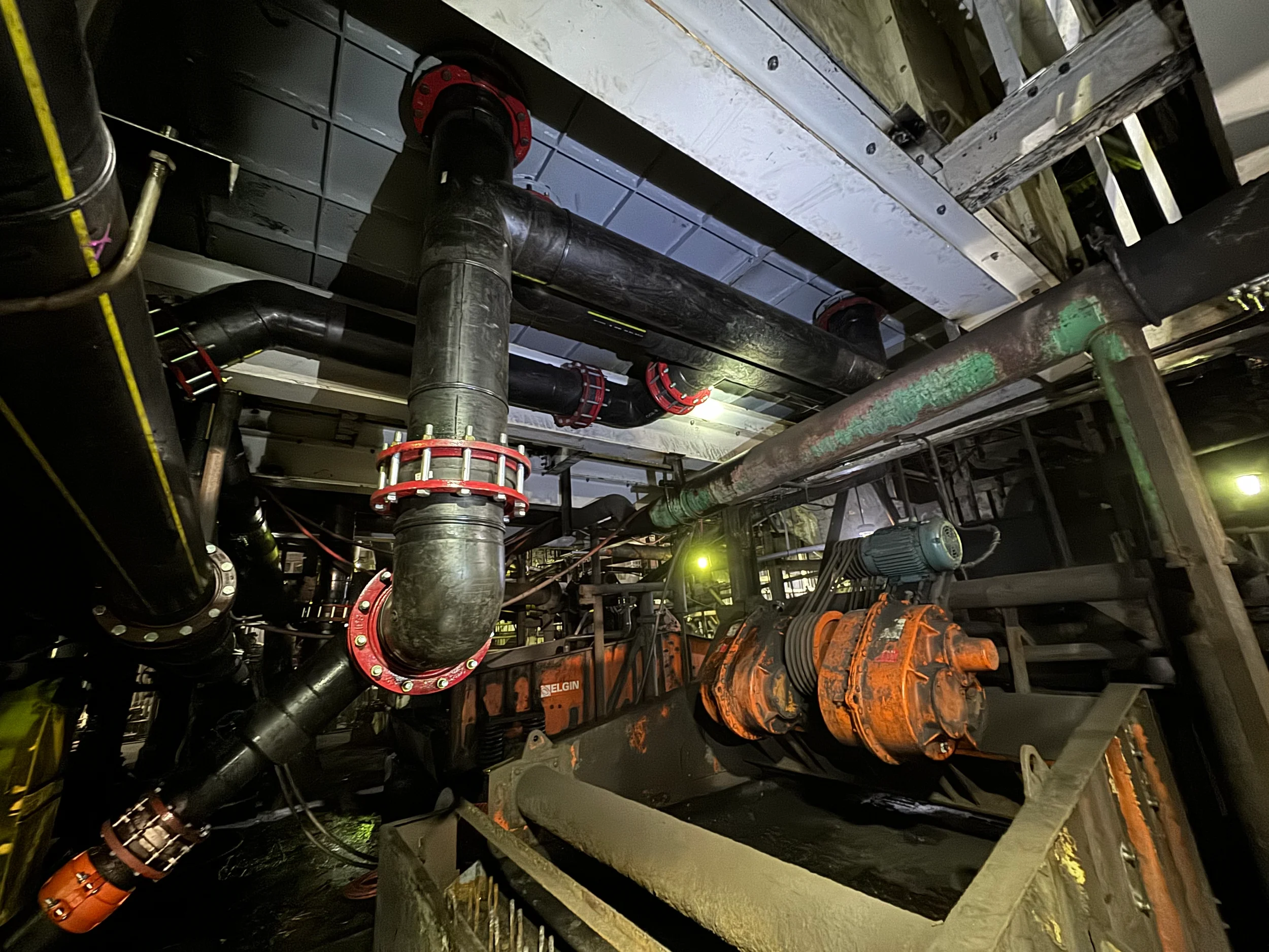 Underground construction site with large black pipes, some with red flanges, connected throughout. Metal scaffolding and equipment are visible, with some pipes painted green and orange. The area appears industrial, with a ceiling overhead and constru