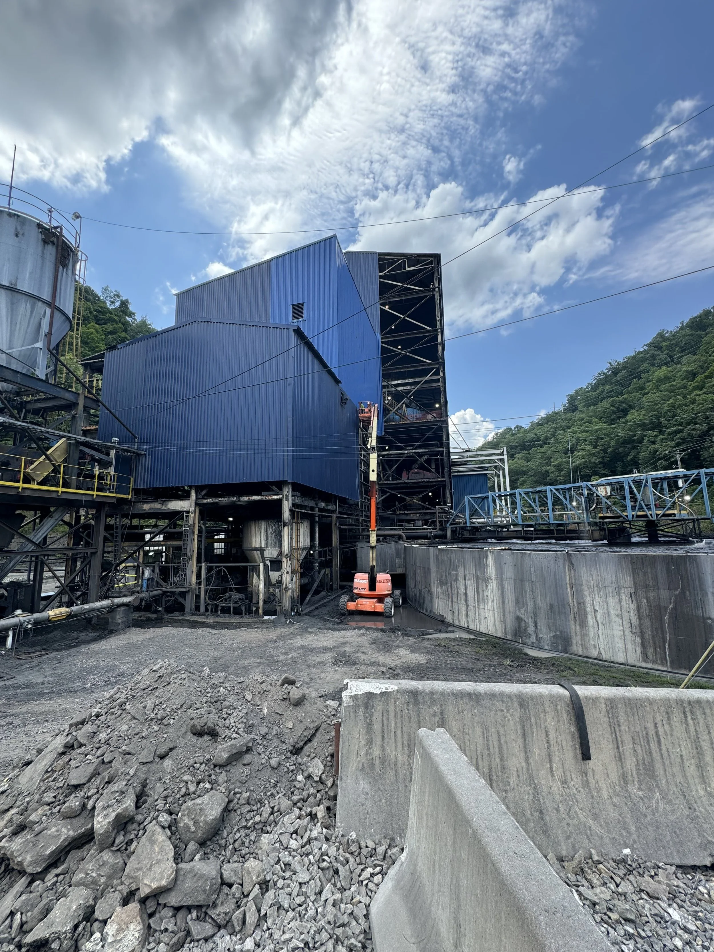 Construction site with blue and black metal structures, a pile of grey rubble, and a large concrete barrier in the foreground. The sky is partly cloudy with scattered clouds.