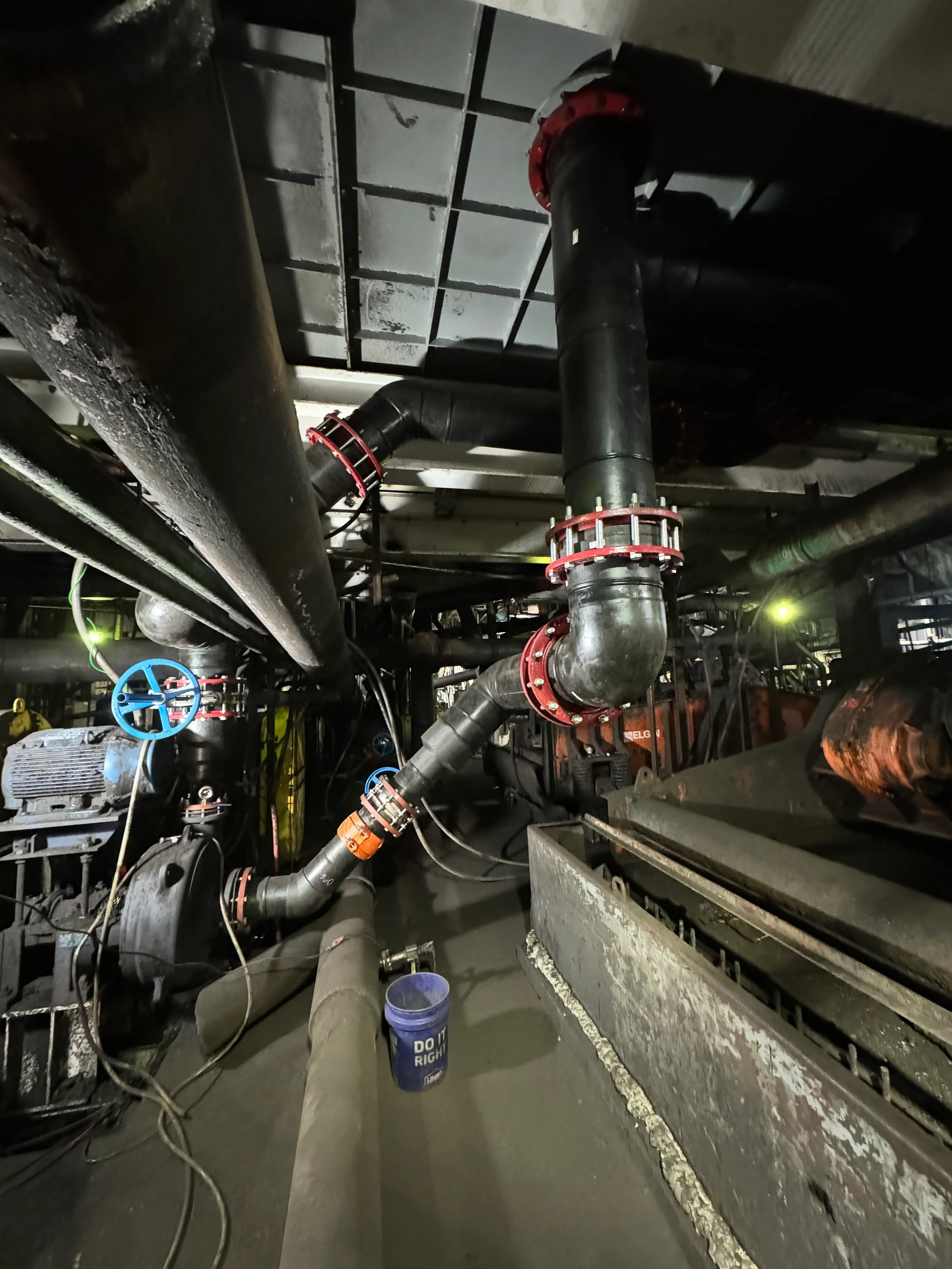Underground mechanical room with black pipes, blue valve handles, cables, and a bucket on the floor.