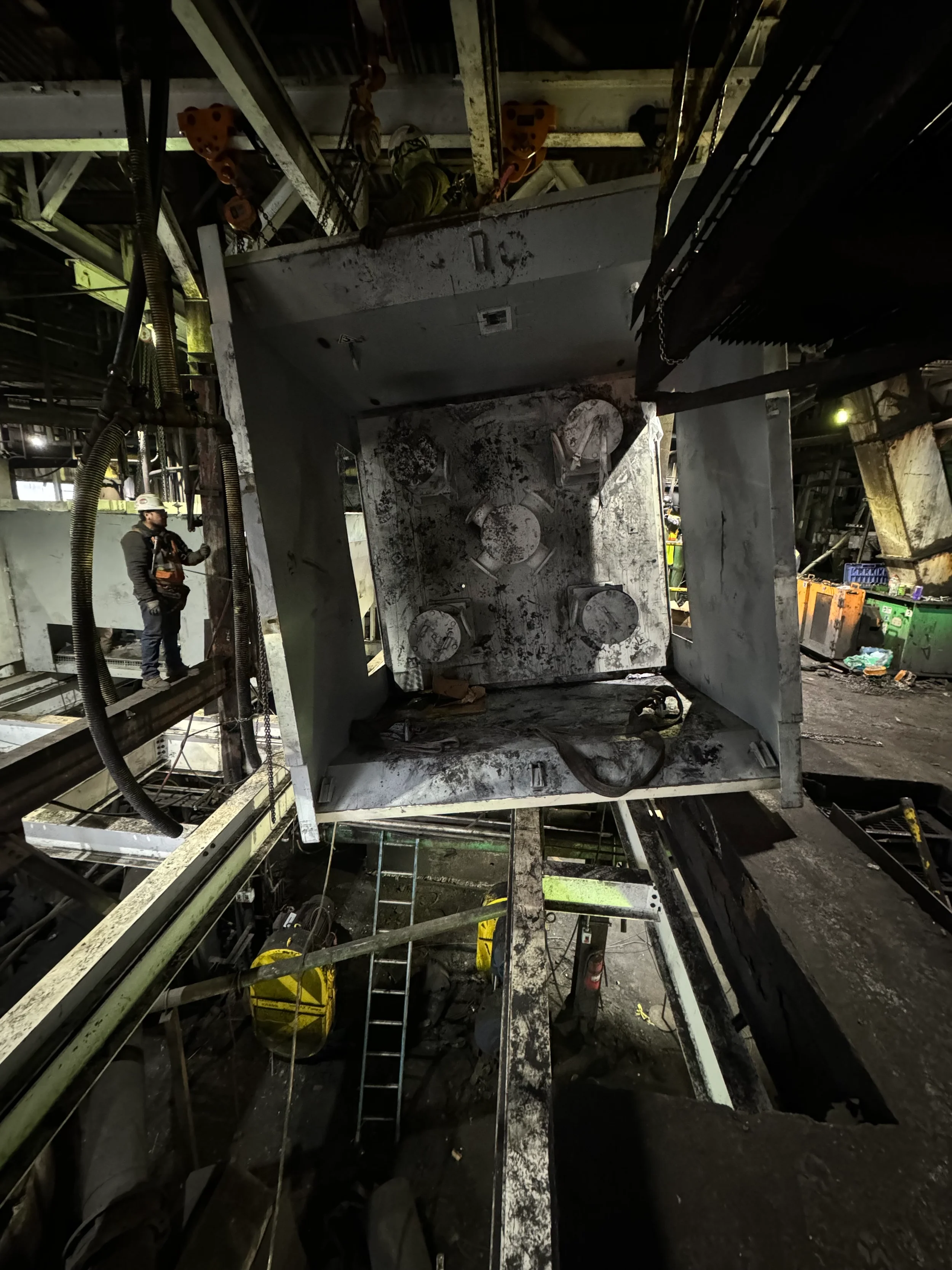 A construction worker wearing safety gear and helmet standing among large industrial equipment with dirty, worn surfaces in a factory or construction site.