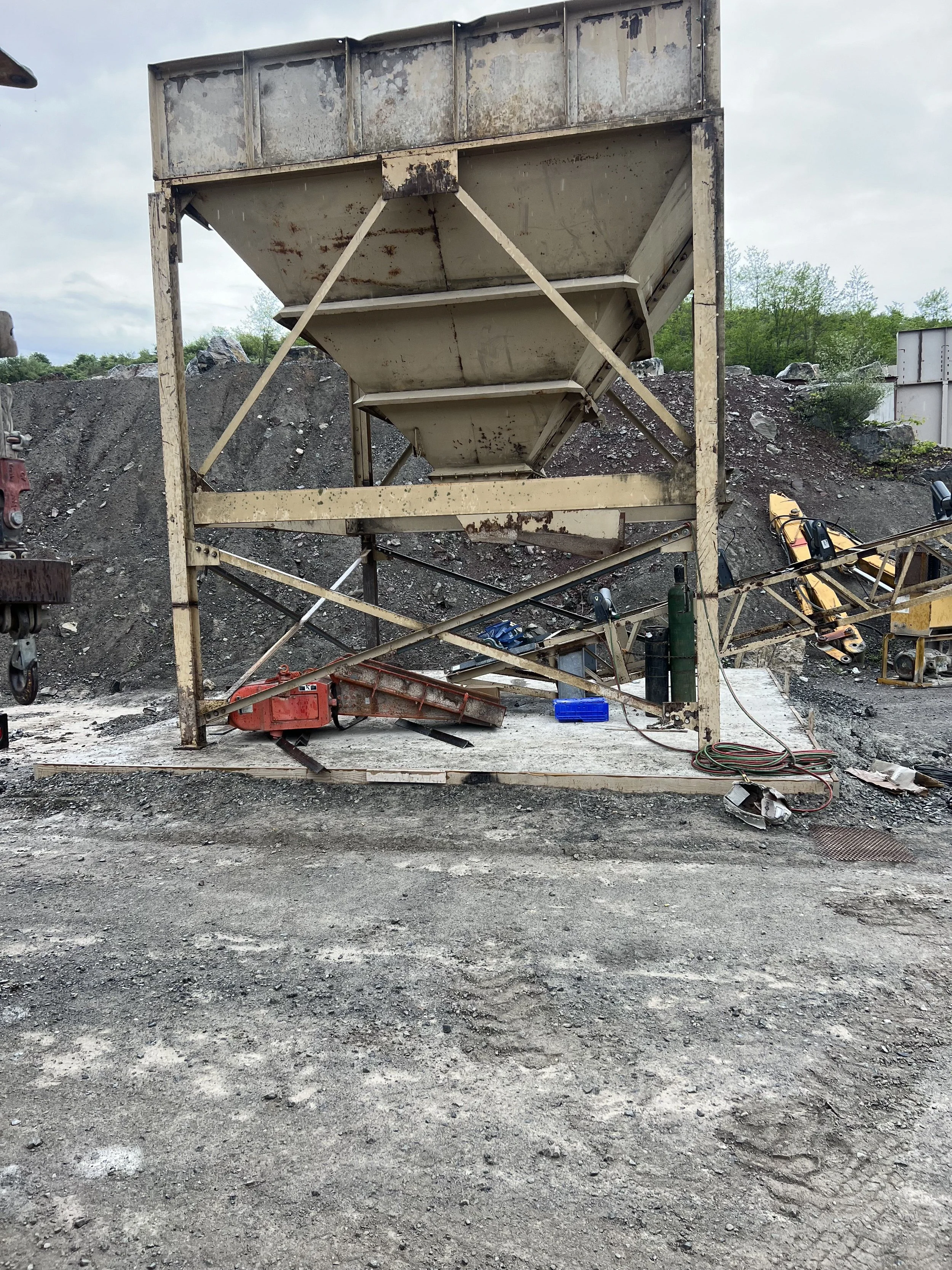 A rusty industrial hopper or chute structure on a construction site with tools, cords, and equipment scattered on the ground in front of it. Gravel and rocks on a hill in the background.
