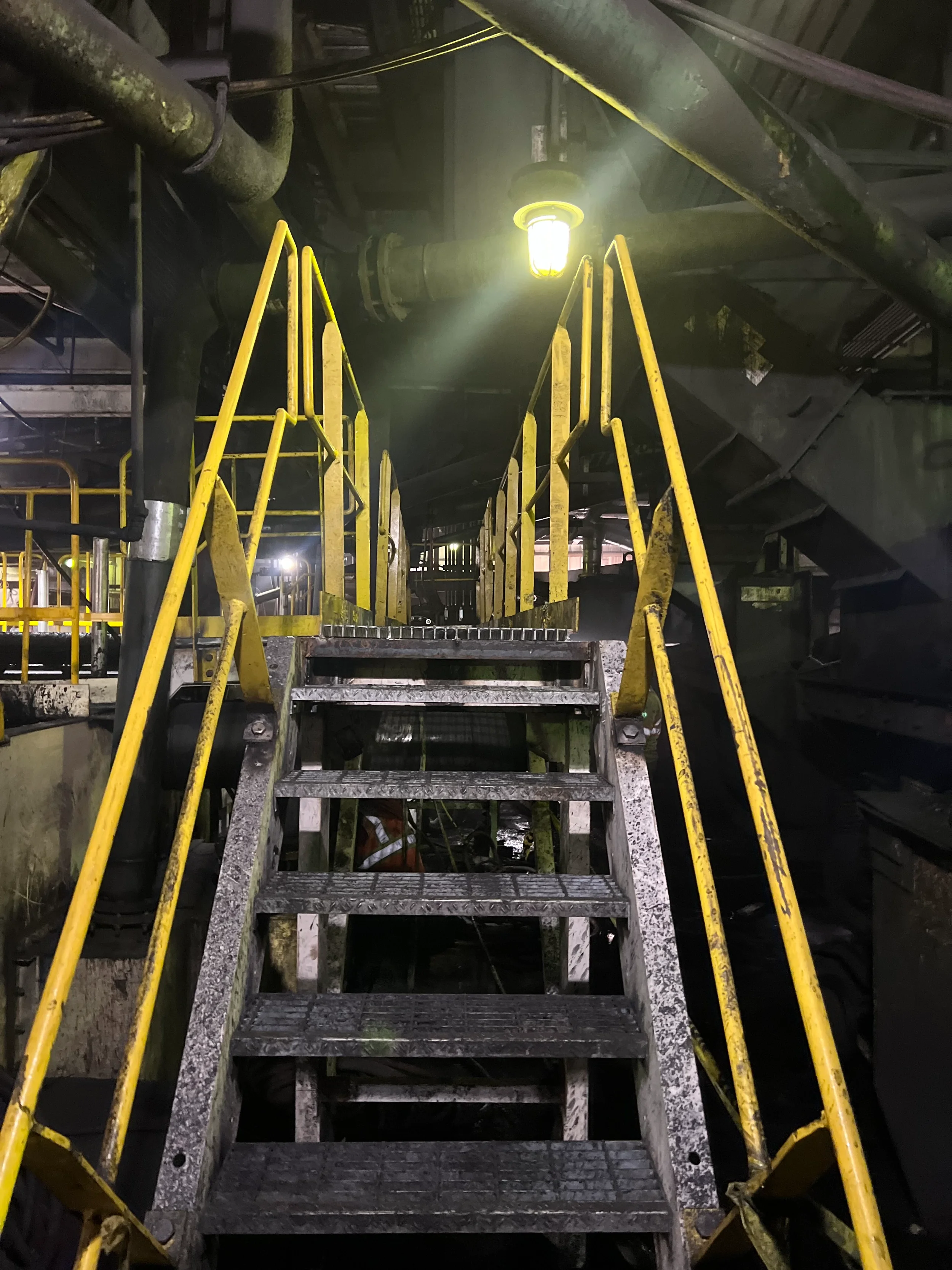 A metal staircase with yellow handrails leading up to a platform inside a large industrial facility. There are large pipes and industrial equipment overhead, and a bright light casts a glow in the background.