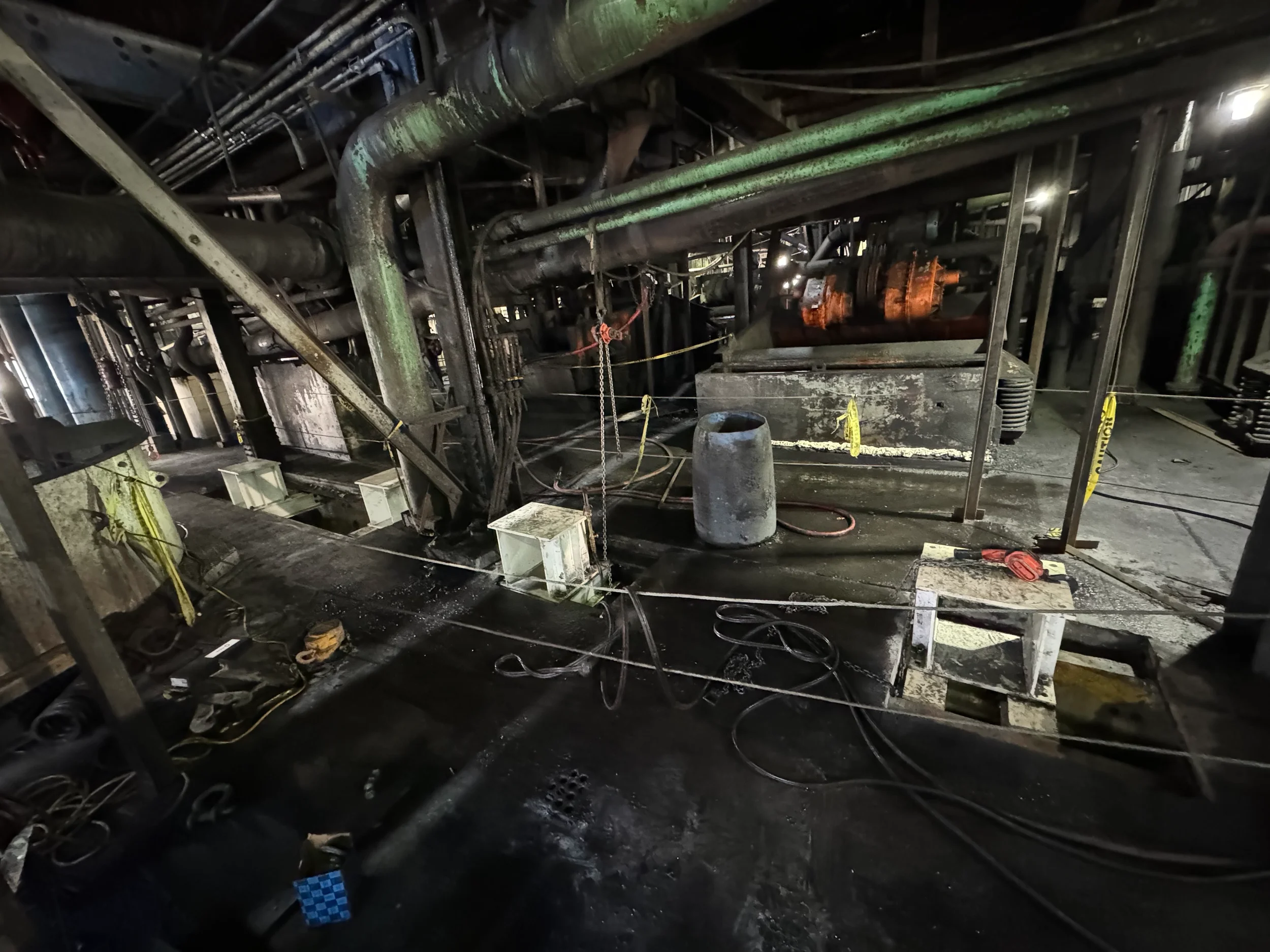 Interior view of a damaged commercial kitchen or industrial facility with burnt and charred equipment, pipes, and debris scattered on the floor, indicating fire damage.