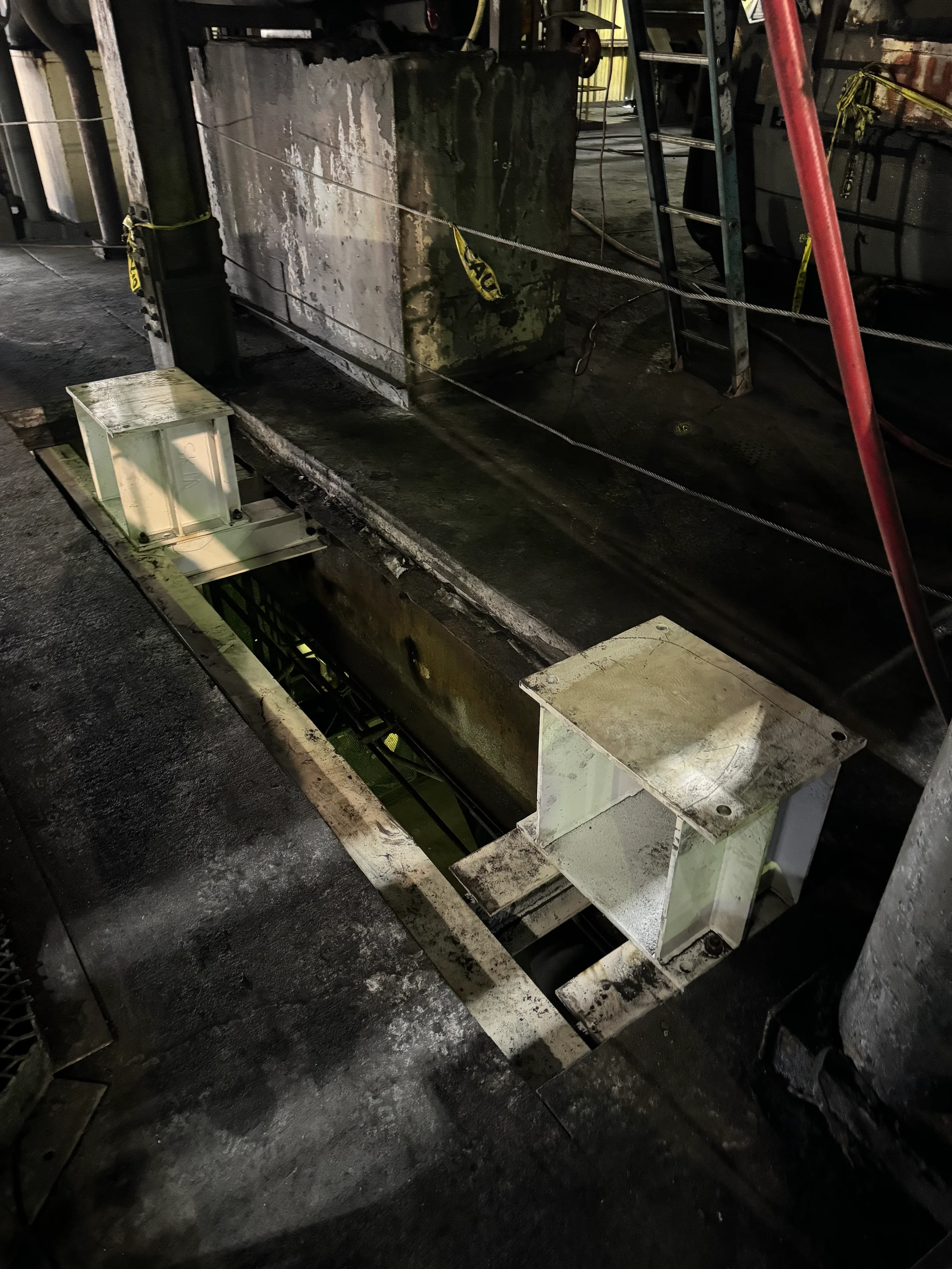 Industrial or construction site with large concrete block, metal framework, ladders, and safety tape, on a dark, worn surface.