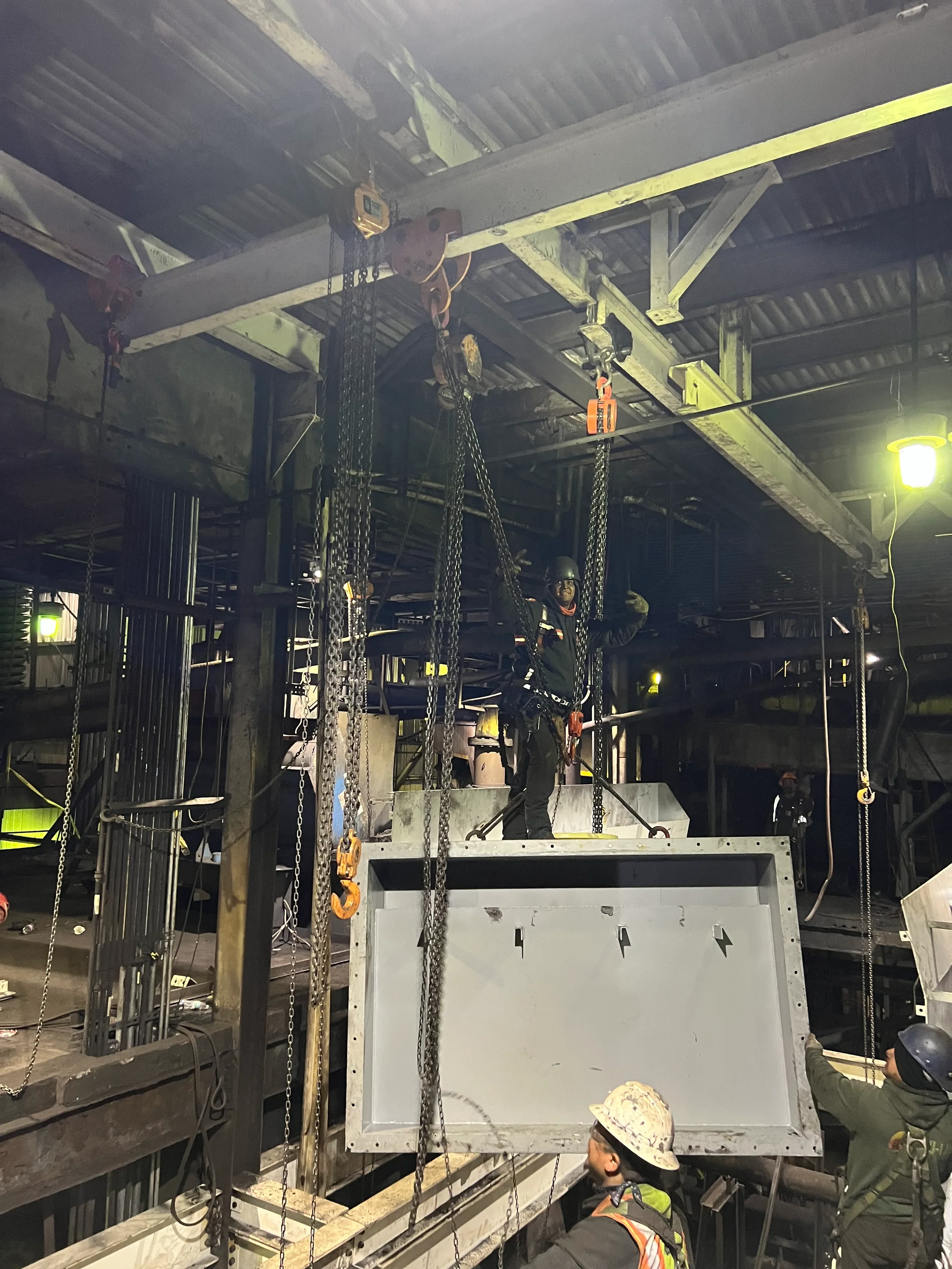 Construction workers wearing safety helmets working inside a building, installing large structural components and using lifting equipment.