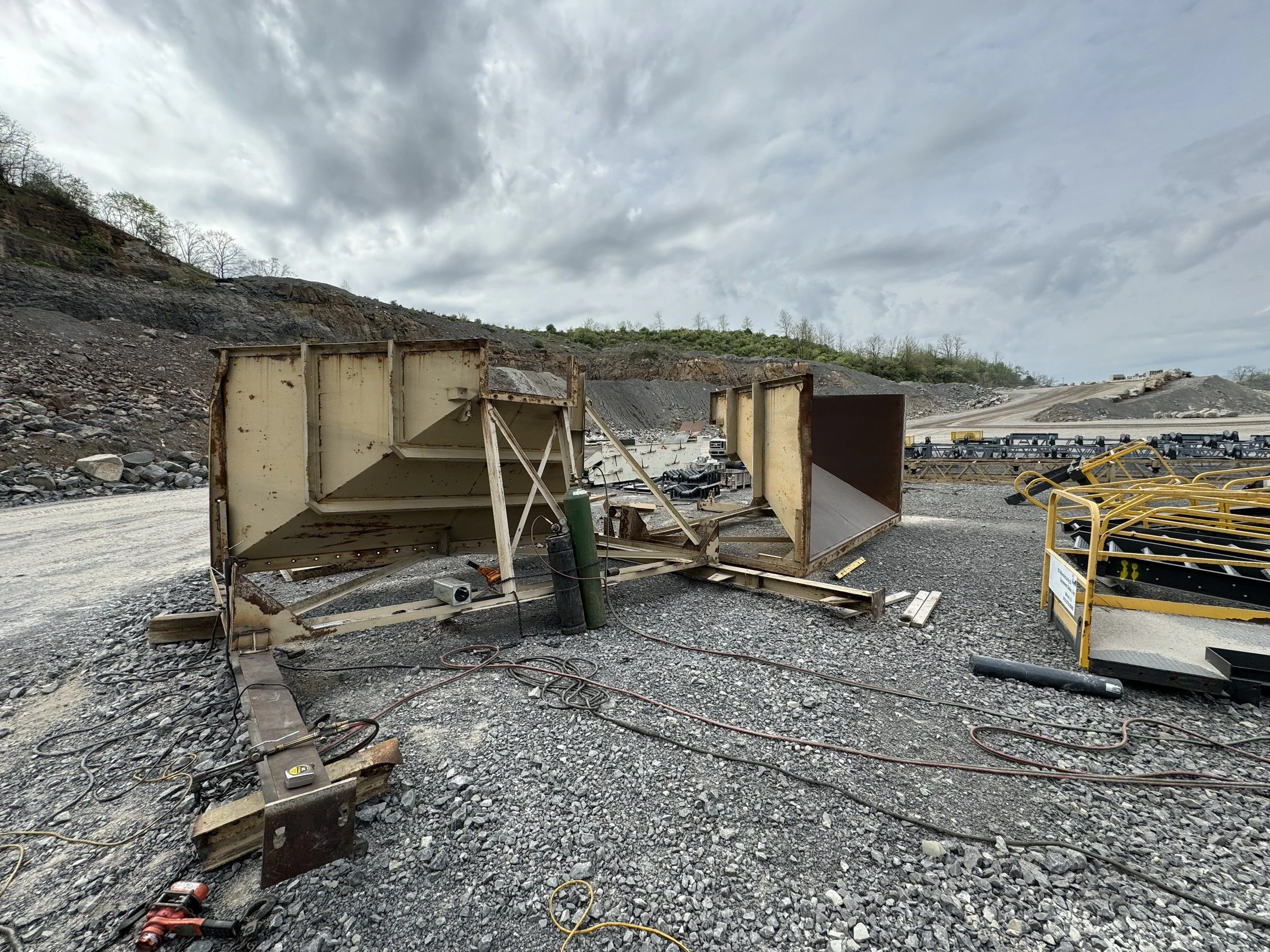 Construction site with remains of rusted metal structures, wires, and equipment, on gravel ground, with a hillside and cloudy sky in the background.