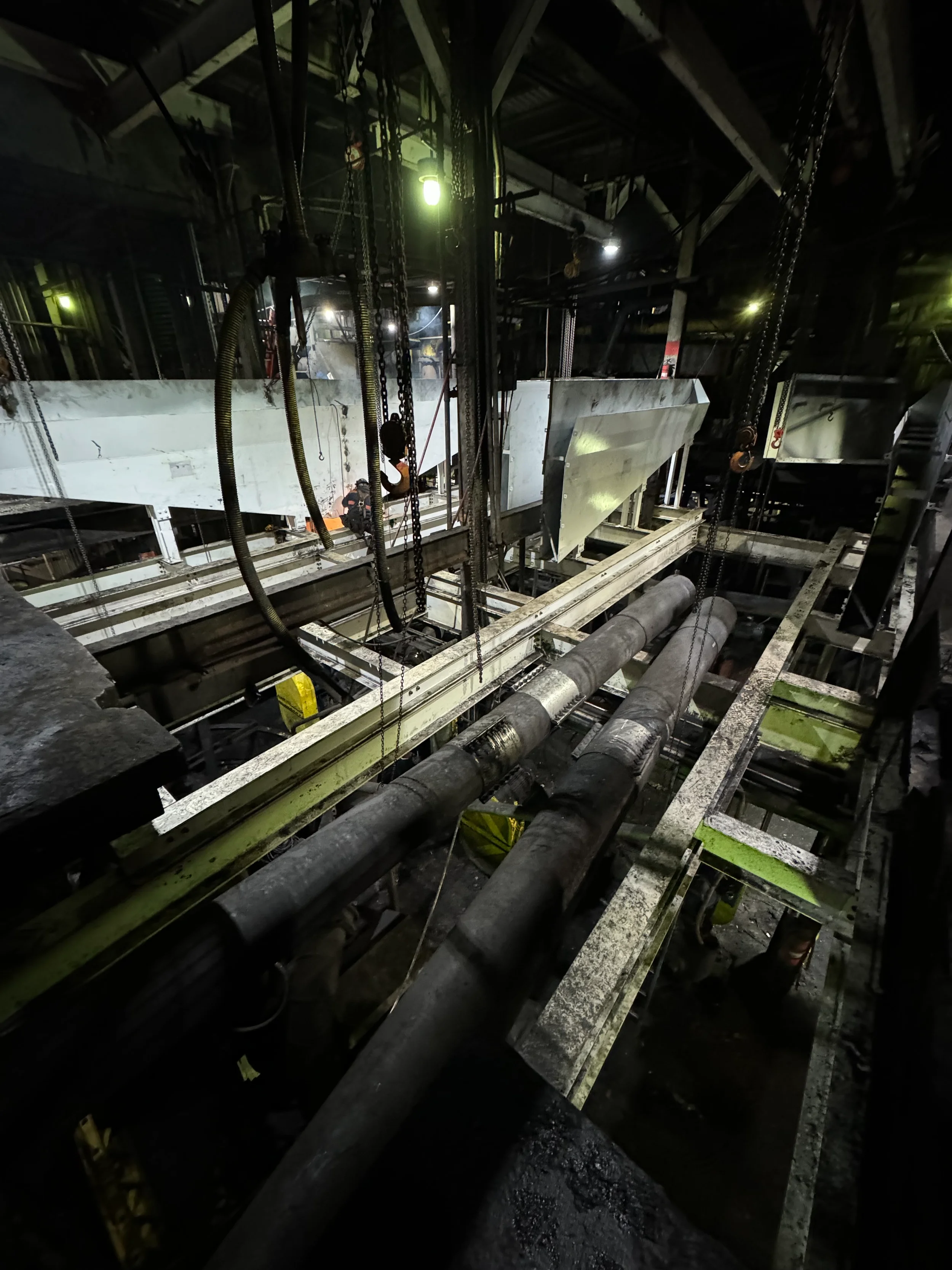 A dark underground construction site featuring metal beams, pipes, and safety chains hanging from the ceiling, with a worker wearing safety gear and a headlamp working on a concrete structure.