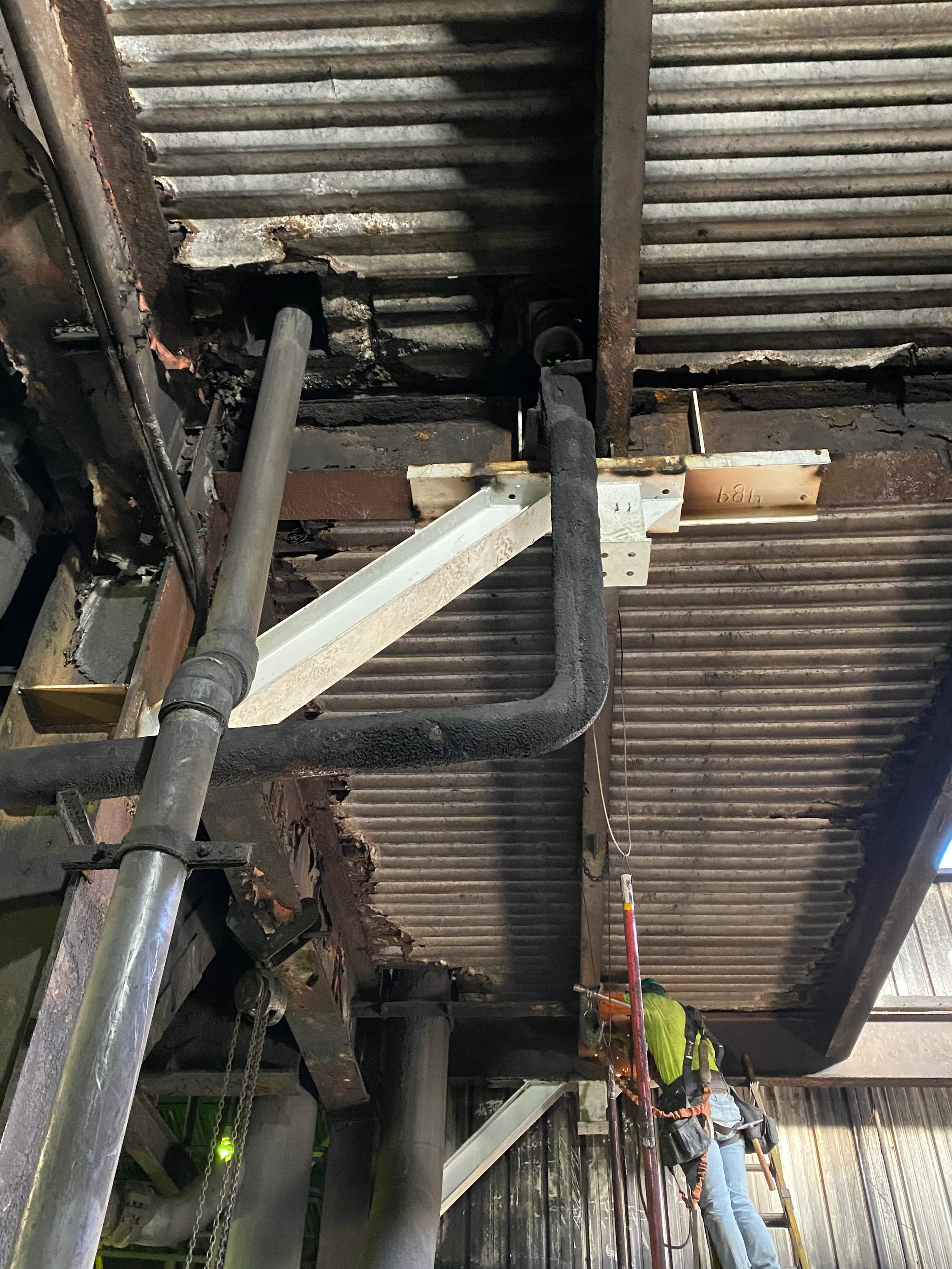 Workers in safety gear repairing or inspecting a damaged ceiling with exposed corrugated metal panels, supported by scaffolding and ladders.