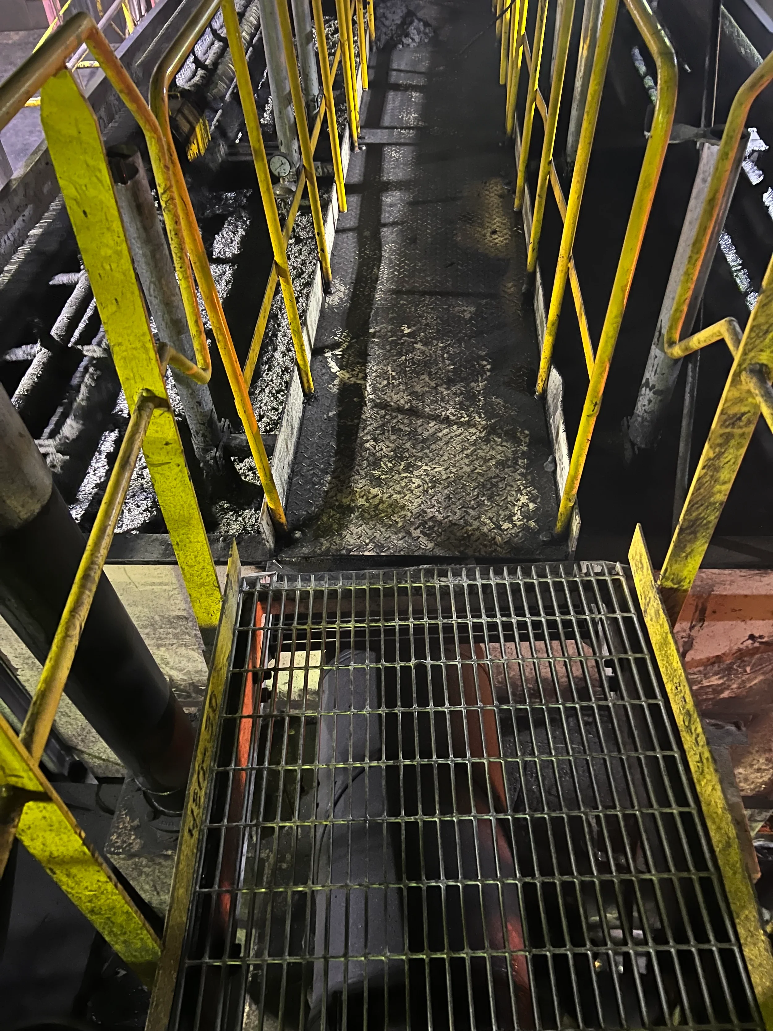 A metal catwalk with yellow safety railings on both sides, leading to a dark area with blackened surfaces and visible industrial pipes.