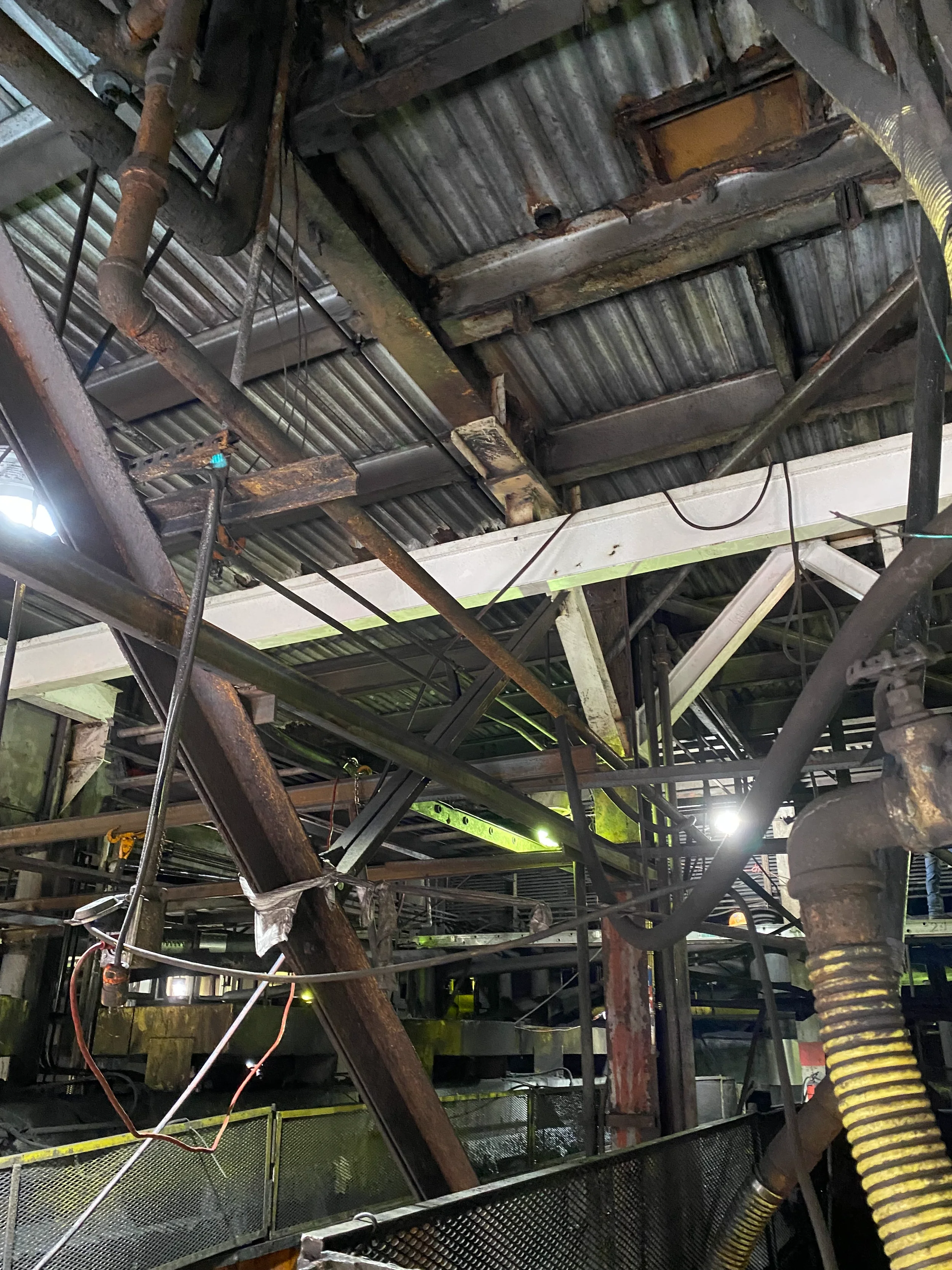 View of the underside of a structure with rusted metal beams, pipes, and a corrugated metal roof. Various wires and hoses are visible, along with a yellow flexible pipe and a metal railing at the bottom. The image shows signs of rust, corrosion, and 